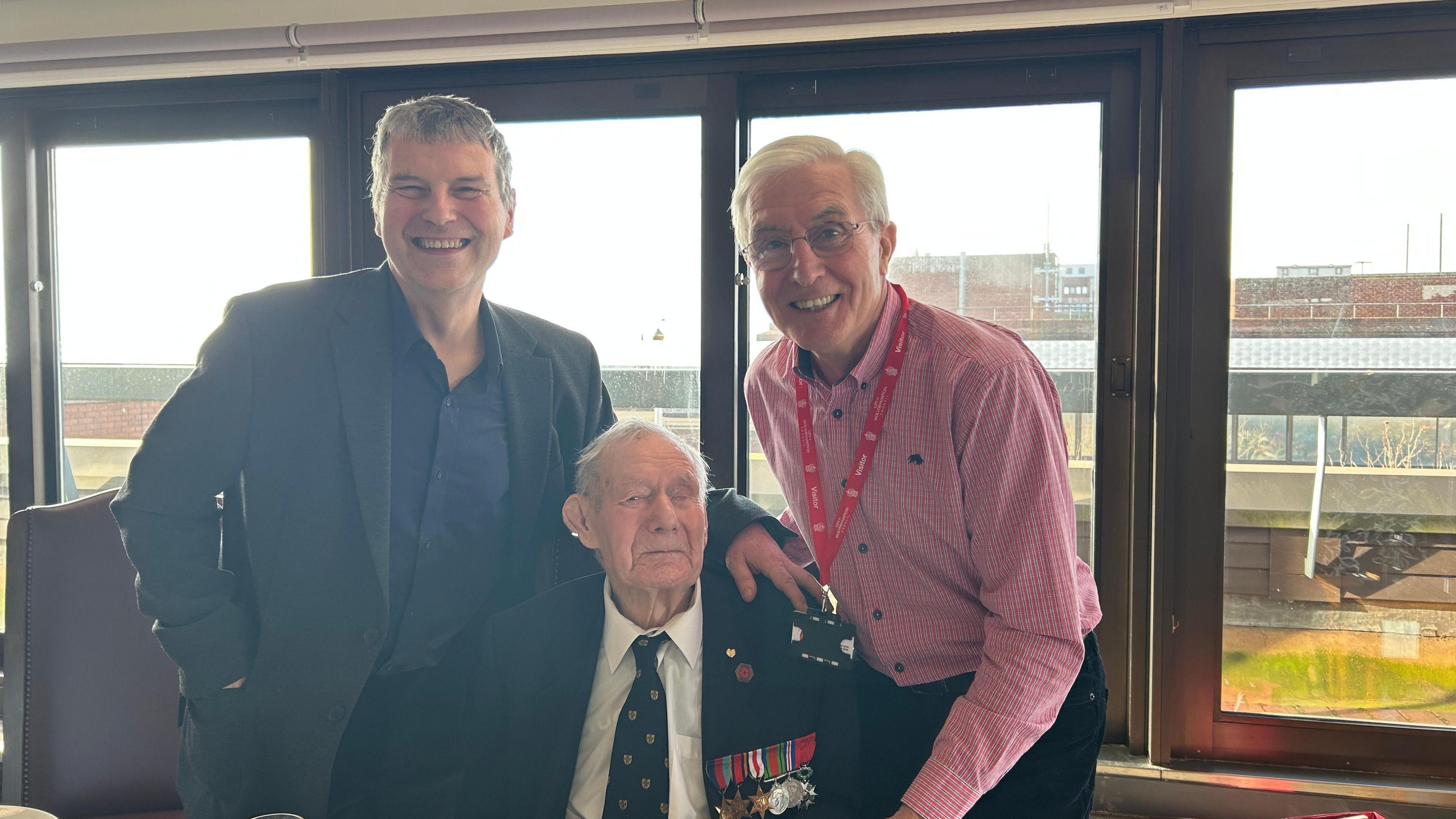 Bill is sat down wearing a dark jacket with his military medals pinned on as well as a white shirt and dark tie. Stood beside him is his son Chris on the left, who wears a dark suit and shirt, and former Wolves player John Richards, who wears a pink shirt, on the right. They stand in front of a row of windows.