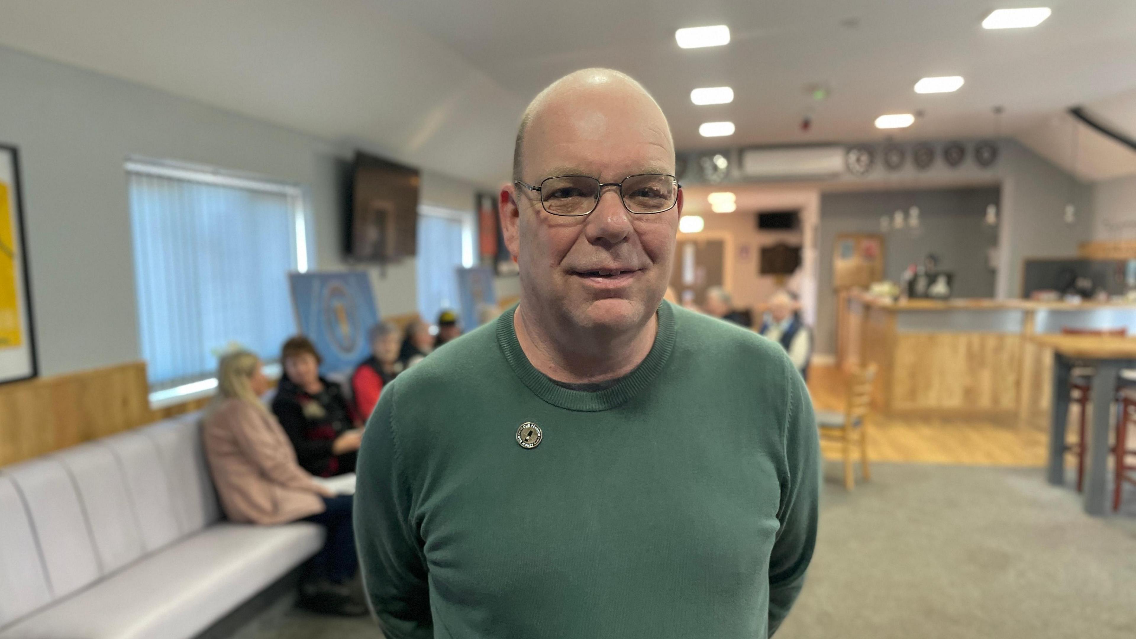 A middle-aged man with bald head and wearing metal-rimmed glasses and green crew-necked top stands in a room with several people seated in the background.