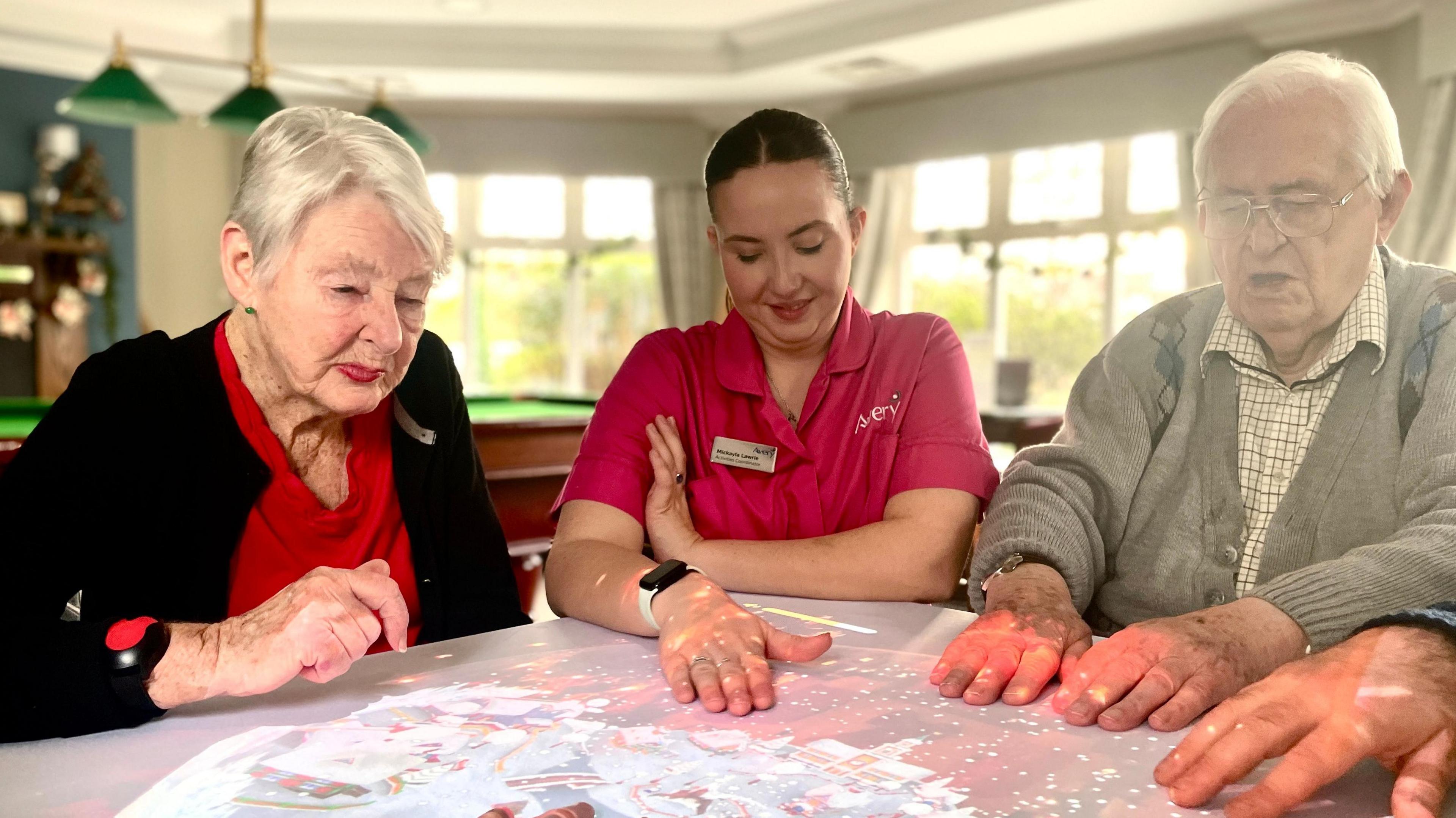 Care home residents and a member of staff sit at a table interacting with  with projected lights on the table.