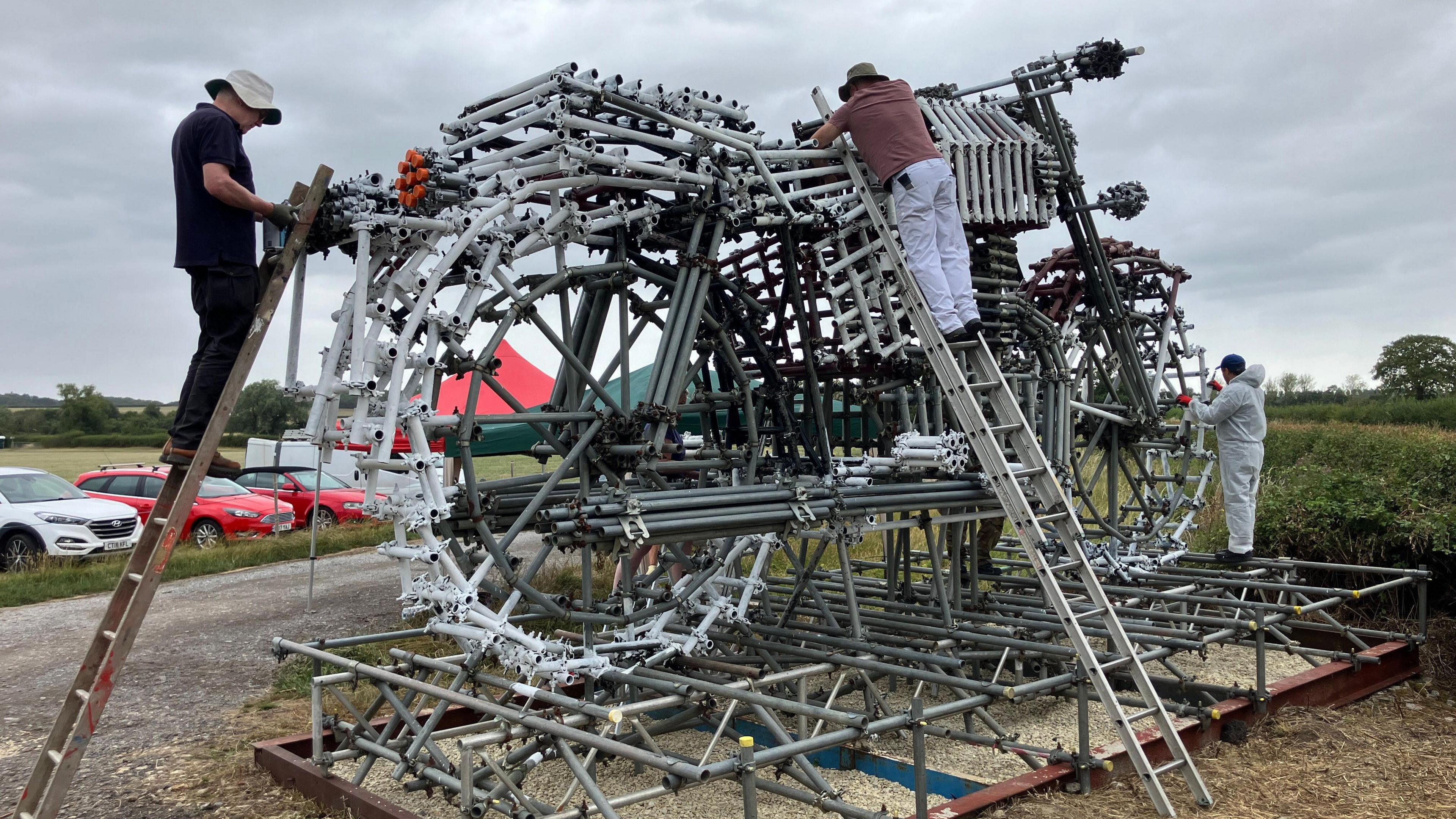 A large sculpture of a motorbike made out of scaffolding poles. There are three men working on the sculpture. Two of them are on ladders while another is stood on the plinth of the sculpture. 