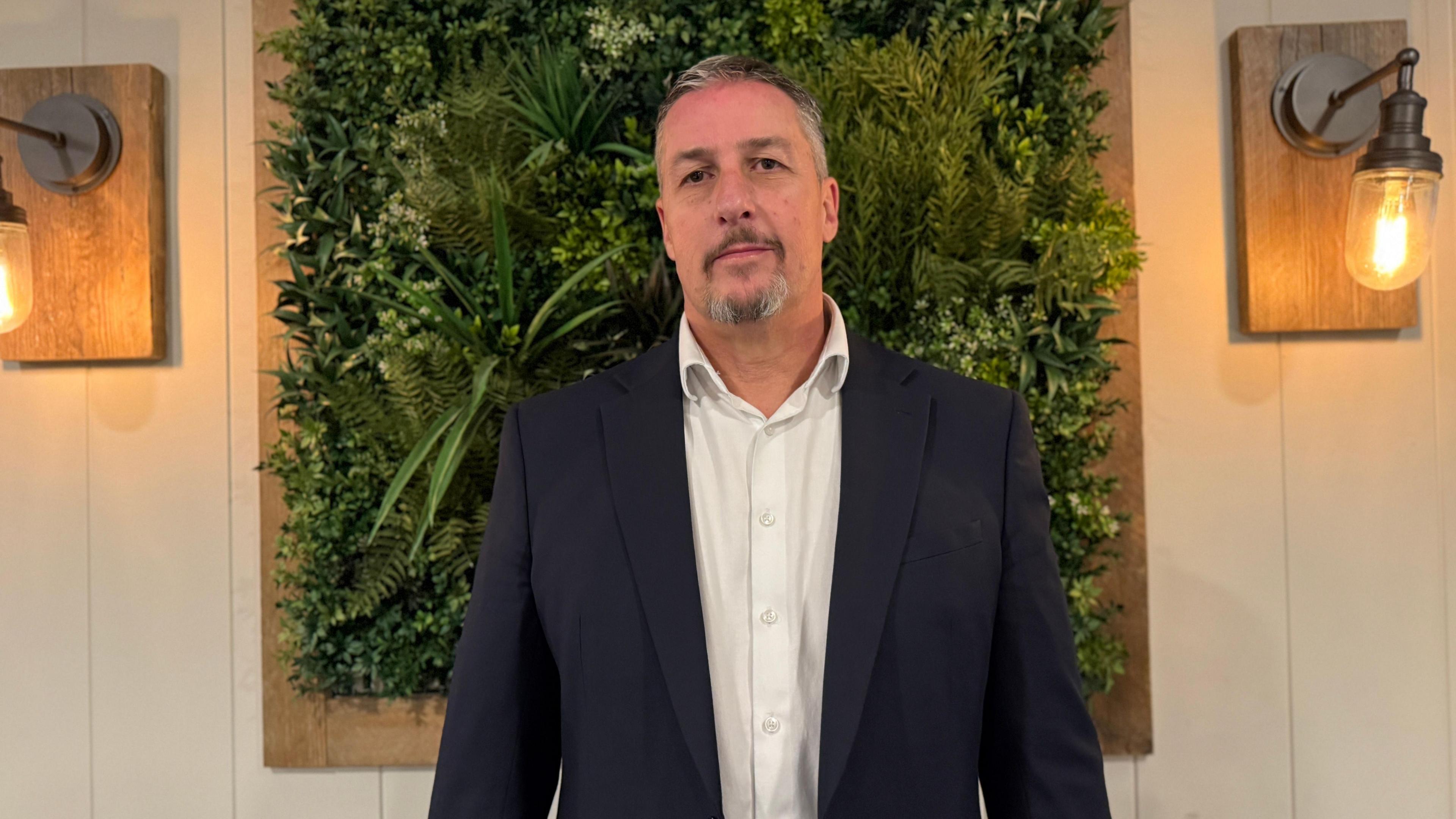 Deputy Andy Cameron, who is a tall middle aged man with short grey hair and a grey goatee beard in a white shirt with the top buttoned opened and a dark navy suit jacket. He is stood in front of a square of fake green plants with wall lights on either side.