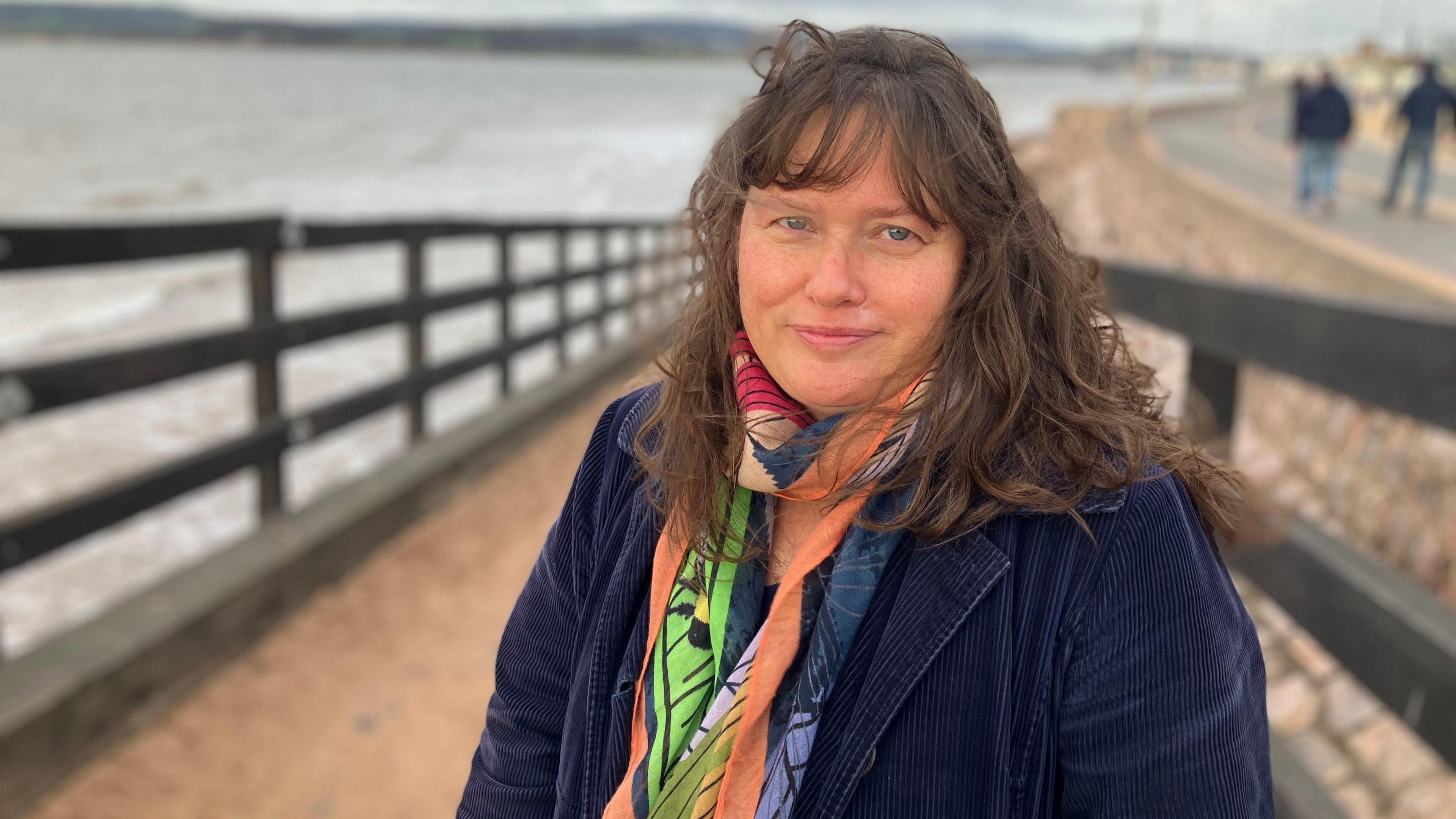 Nicky Nicholls stands on a slipway by Exmouth beach. She has a multi-coloured scarf on and a navy blue corduroy jacket on. She has long brown hair. The tide is in in the background. 