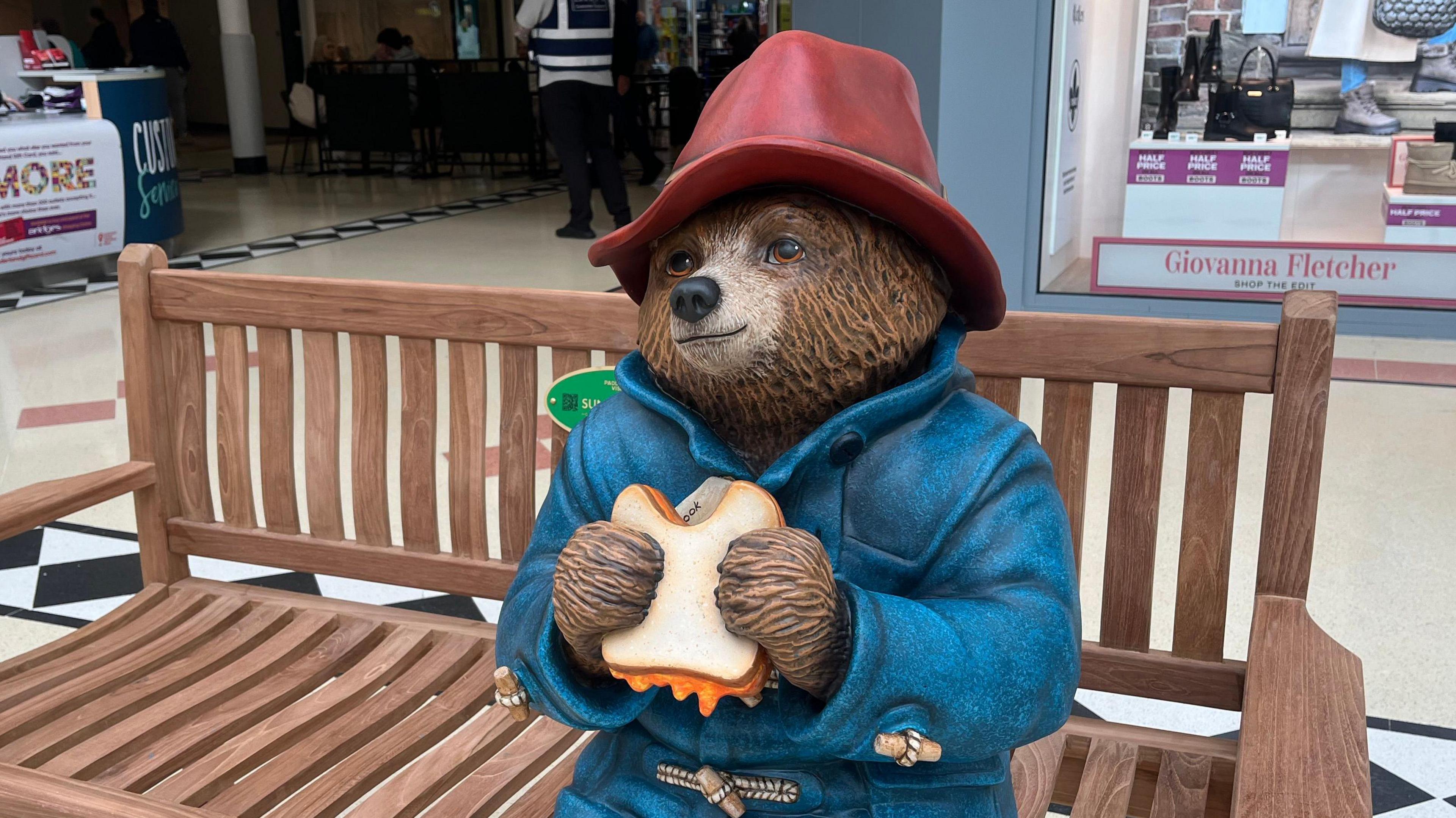 Paddington Bear statue which has been placed in the Bridges shopping centre. Paddington, in red hat and blue coat, sits on a bench eating a marmalade sandwich.