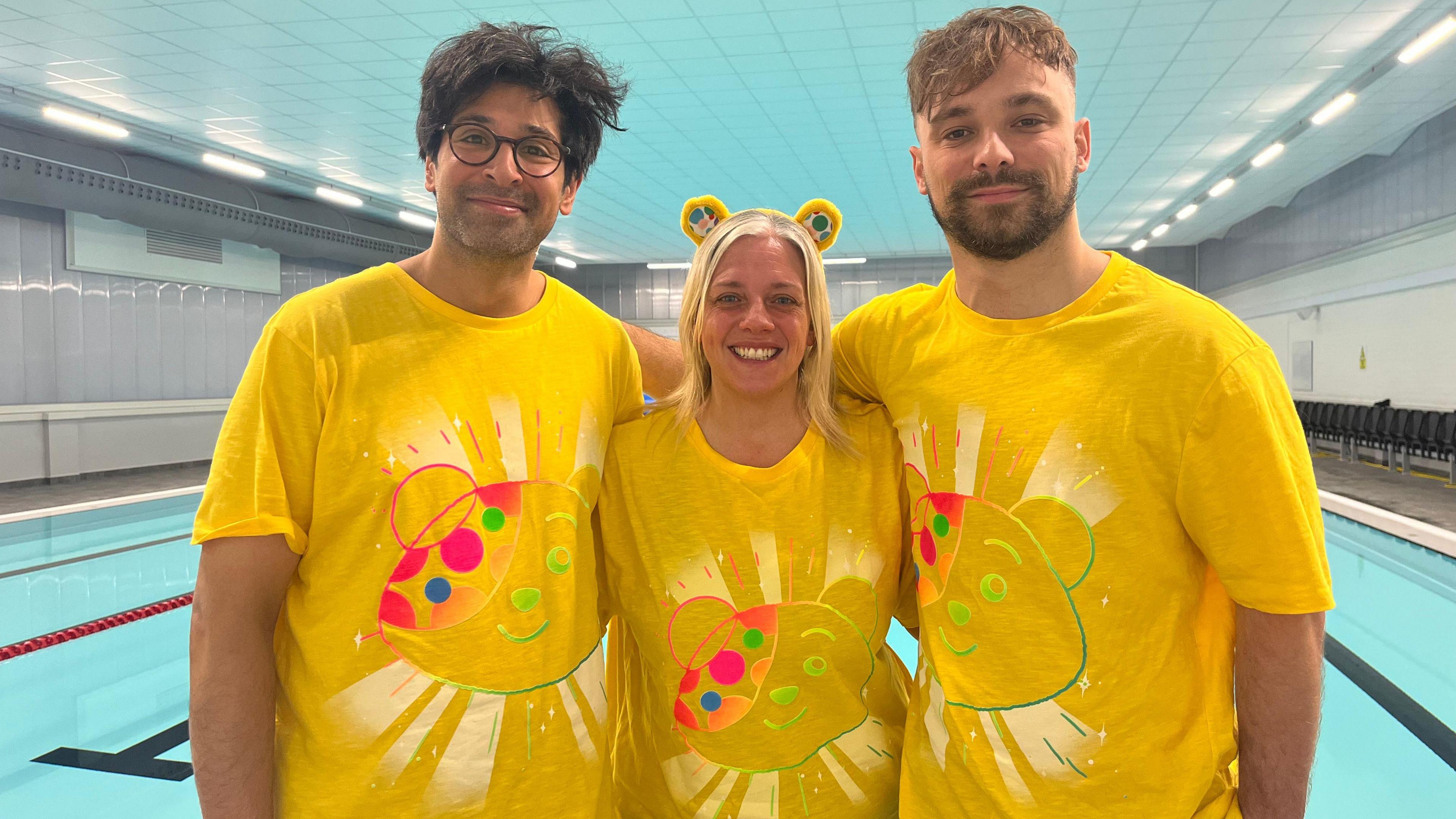 Two men and one woman wearing yellow Children in Need shirts standing next to a swimming pool.