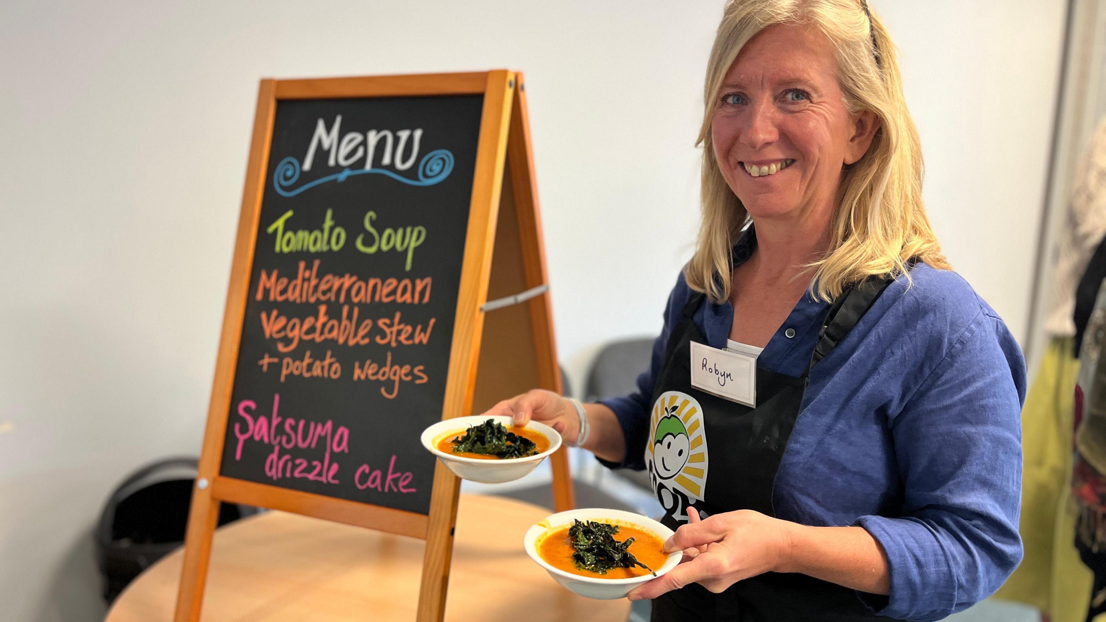 Robyn Creighton holds two bowls of soup with kale on the top, next to a board showing the menu.