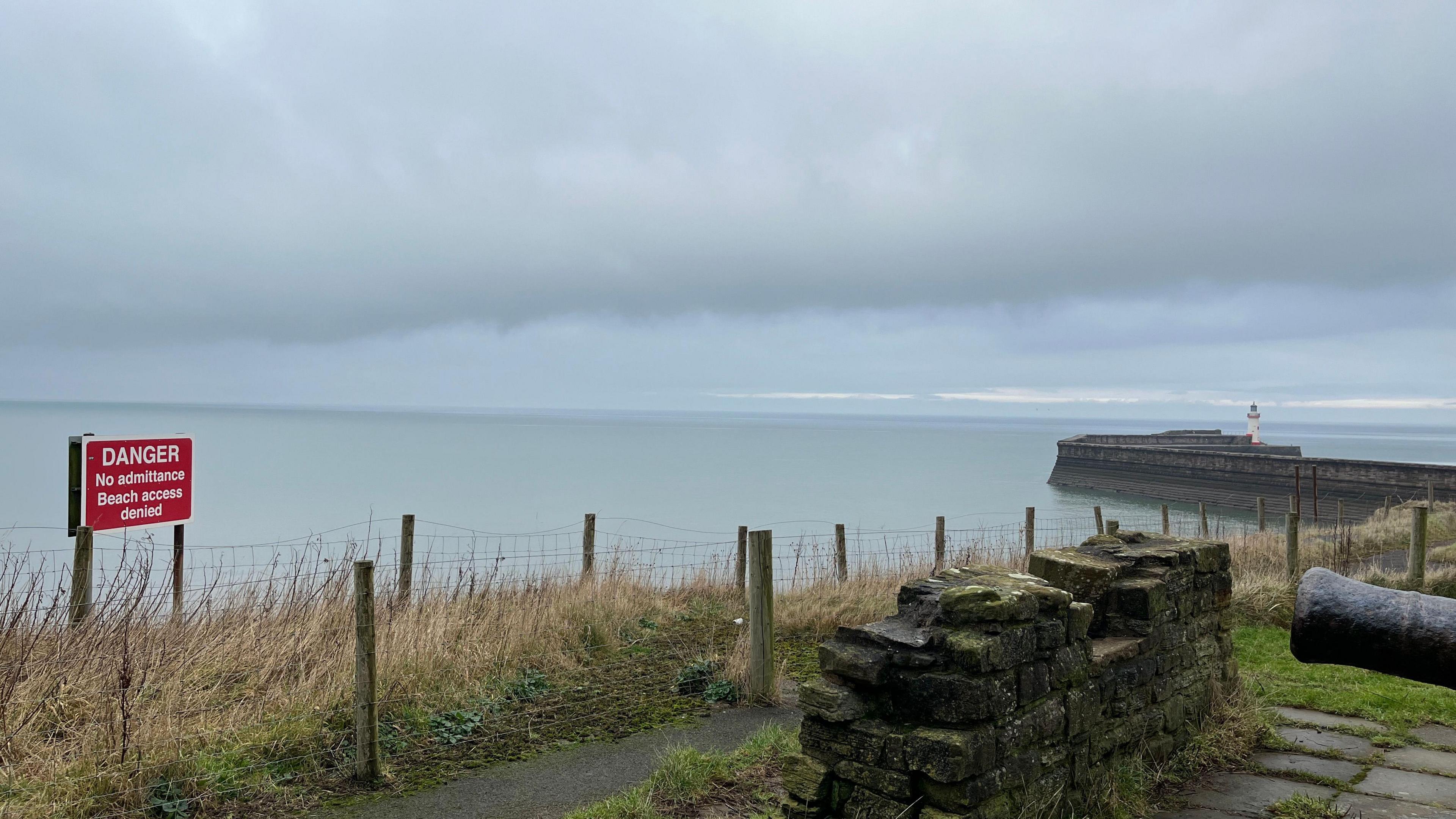 A red sign on the cliff edge saying "Danger, no admittance, beach access denied". The sign is behind a fence. In the background there is the end of the harbour wall with a white and red lighthouse.