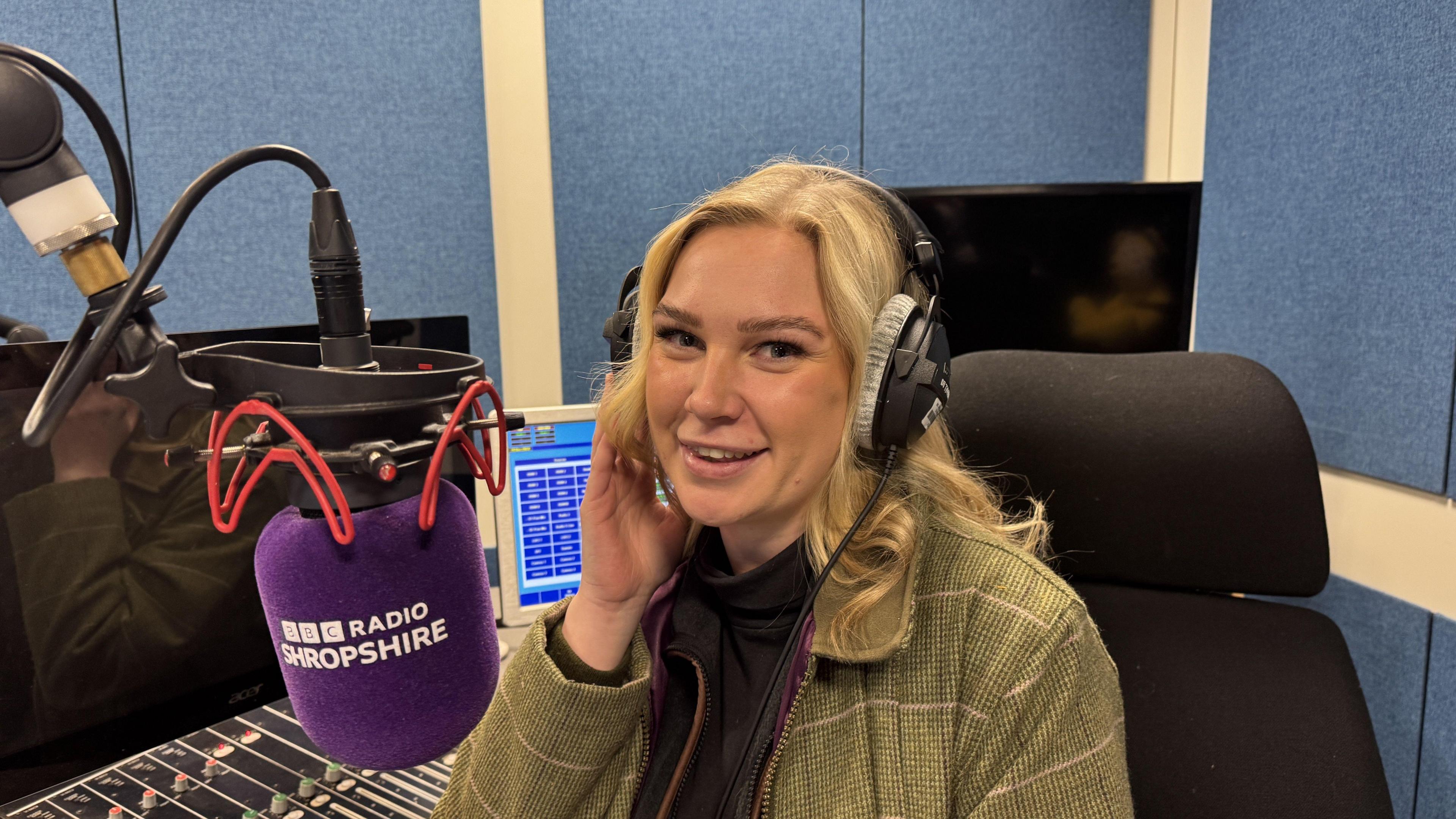 Sarah is sitting in front of a BBC Radio Shropshire purple microphone. She has blonde hair and is wearing headphones. She is wearing a green jacket with purple lining