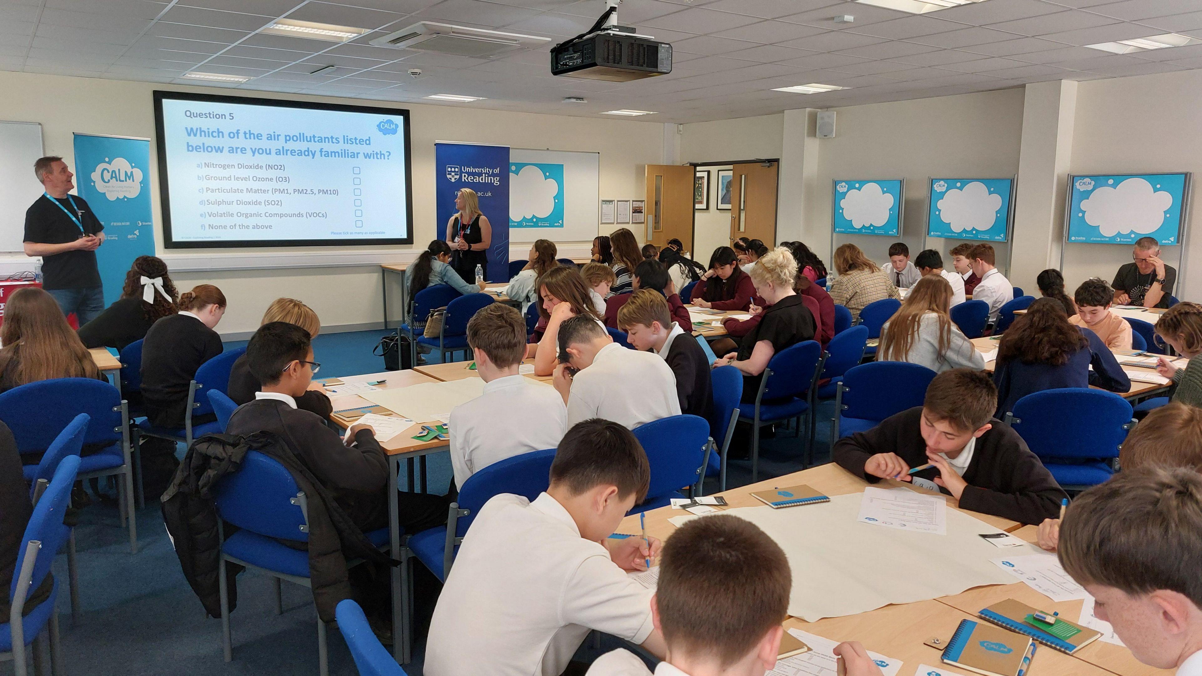 Dozens of students are sat in a classroom. They are wearing black jumpers and white t shirts as their school uniform and all sit around various tables looks at pieces of paper. They are sitting on blue chairs. A man and woman stand at the front of the classroom, either side of a projector screen, talking to the children.