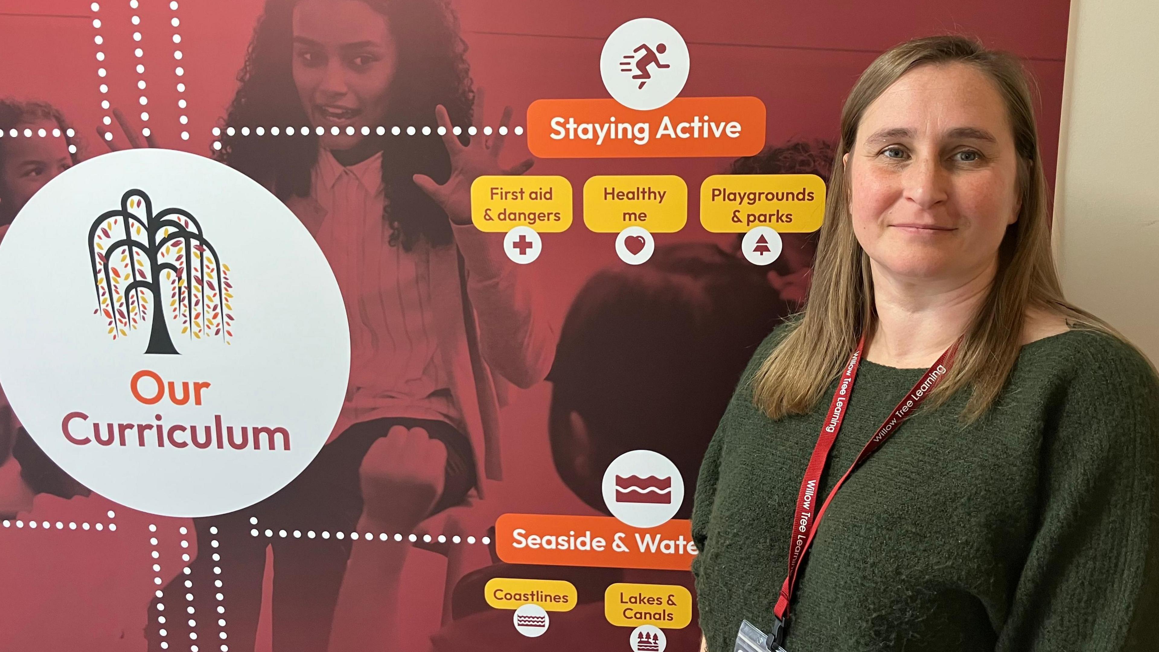 A woman in a green jumper stands next to some red branding on a board, with the Willow Tree Learning logo on it.