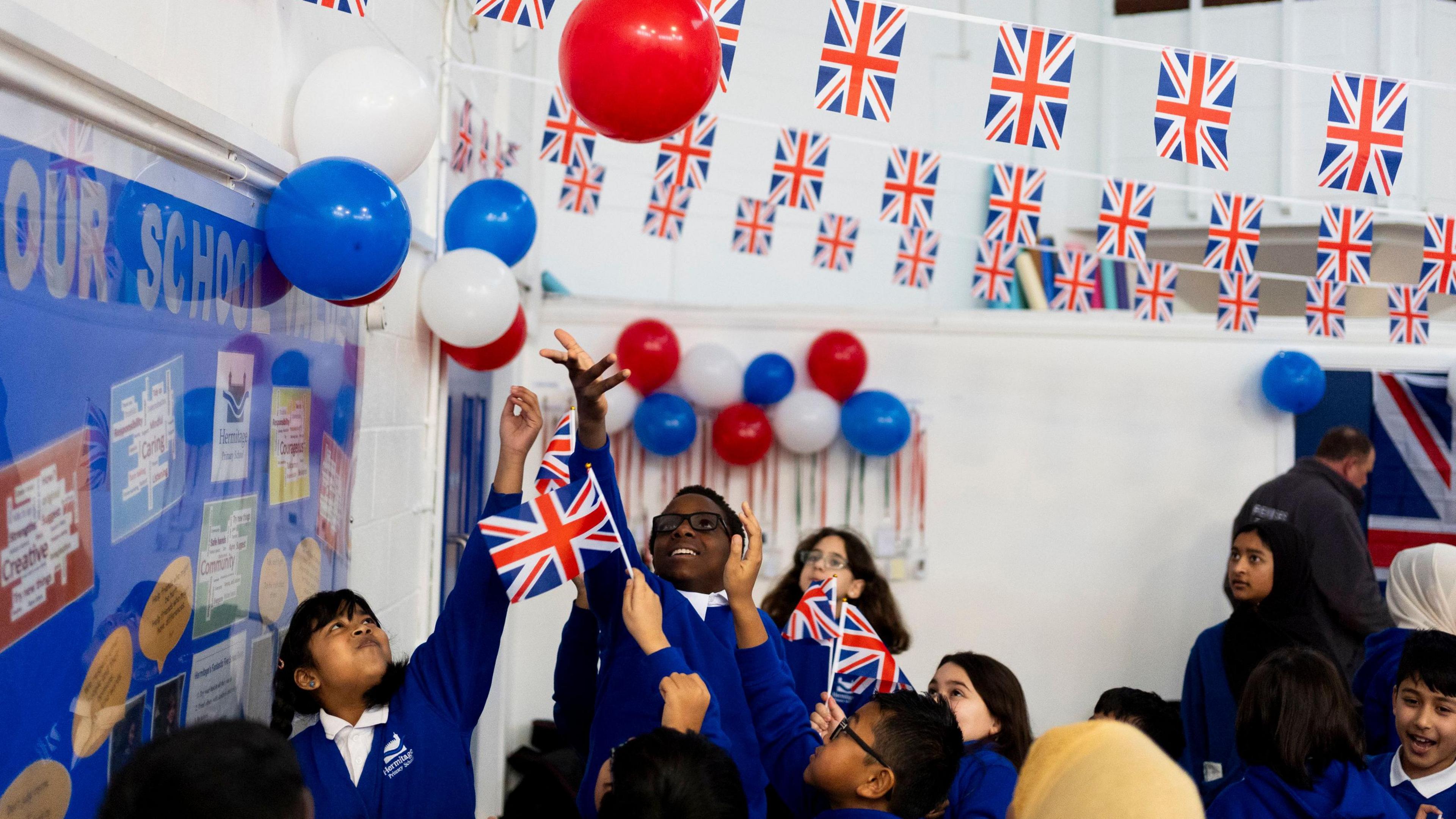 School children dressed in blue reaching up to touch balloons, some holding union flags 