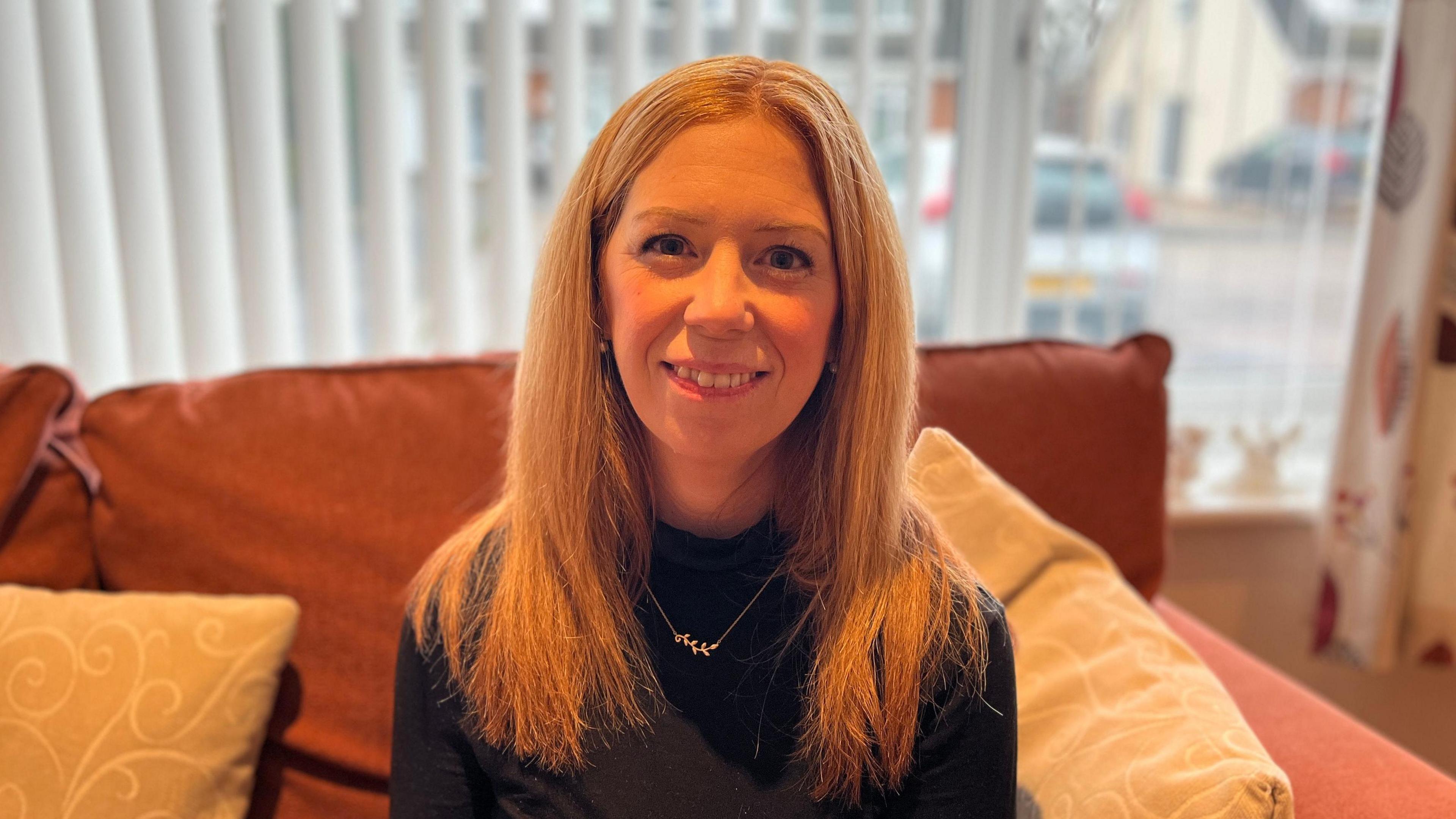 A woman with long ginger hair looking directly into the camera. She is sitting on a sofa in front of a window