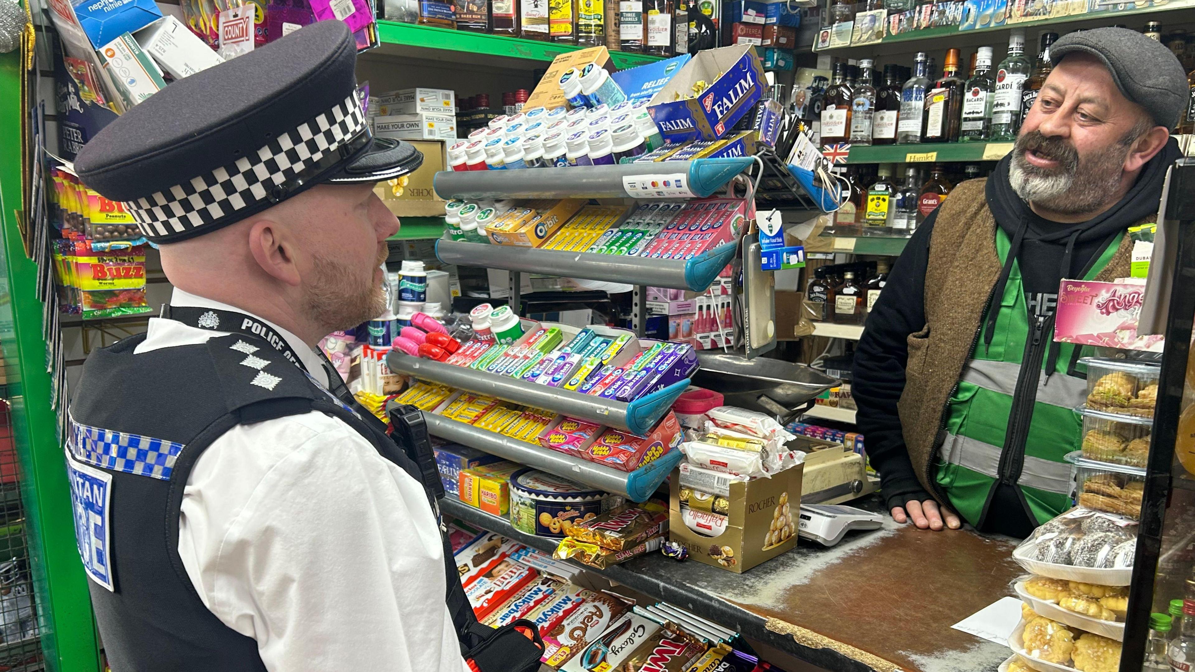 Kryeinspektori në detyrë Charlie Routley ka veshur uniformën e tij të policisë dhe qëndron para banakut të një dyqani duke folur me një punonjës të dyqanit.