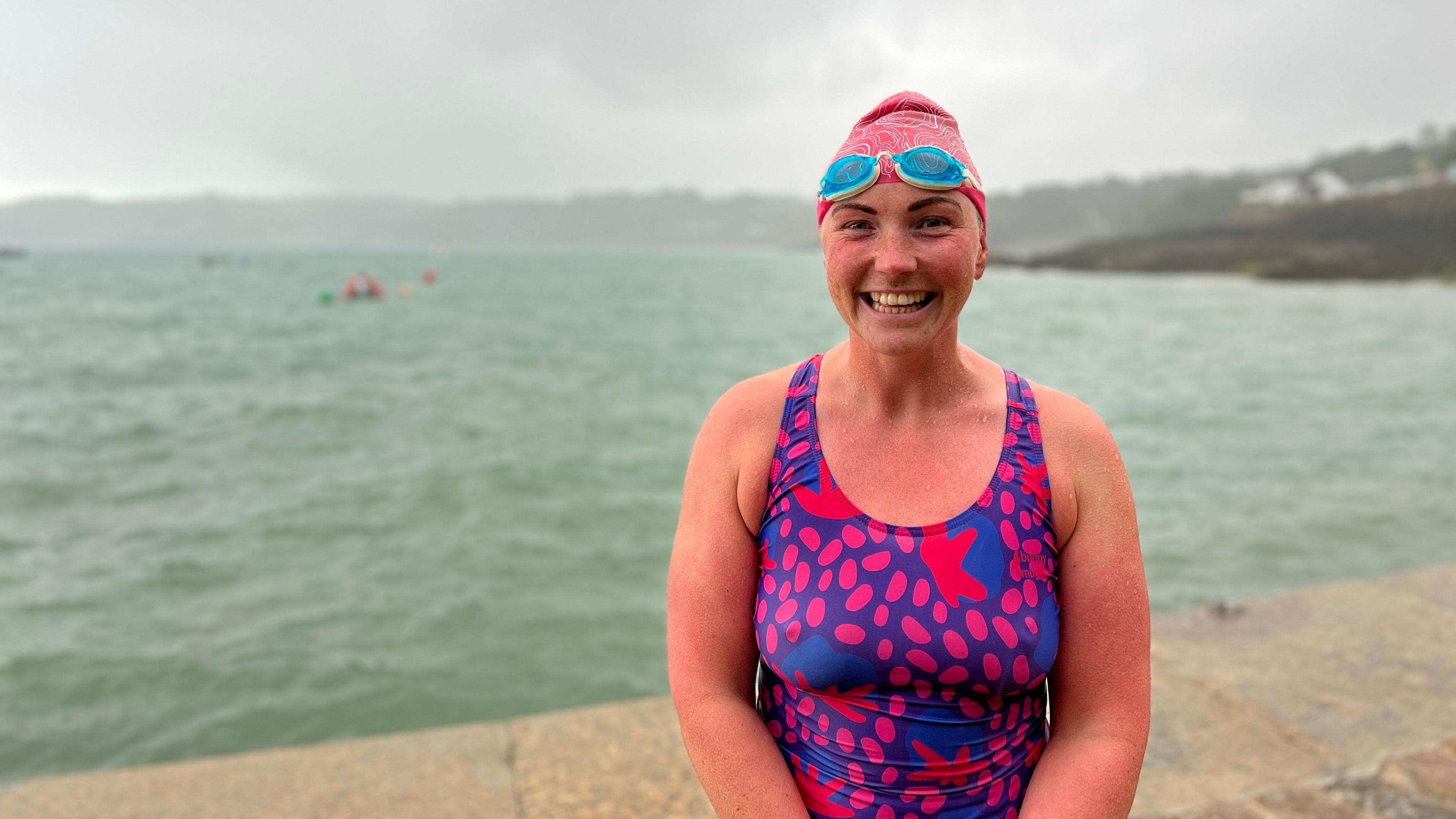 A person in a pink and purple patterned swimsuit and pink swim cap stands on a concrete surface near choppy water, under an overcast sky. Distant swimmers and a shoreline with greenery are visible in the background.