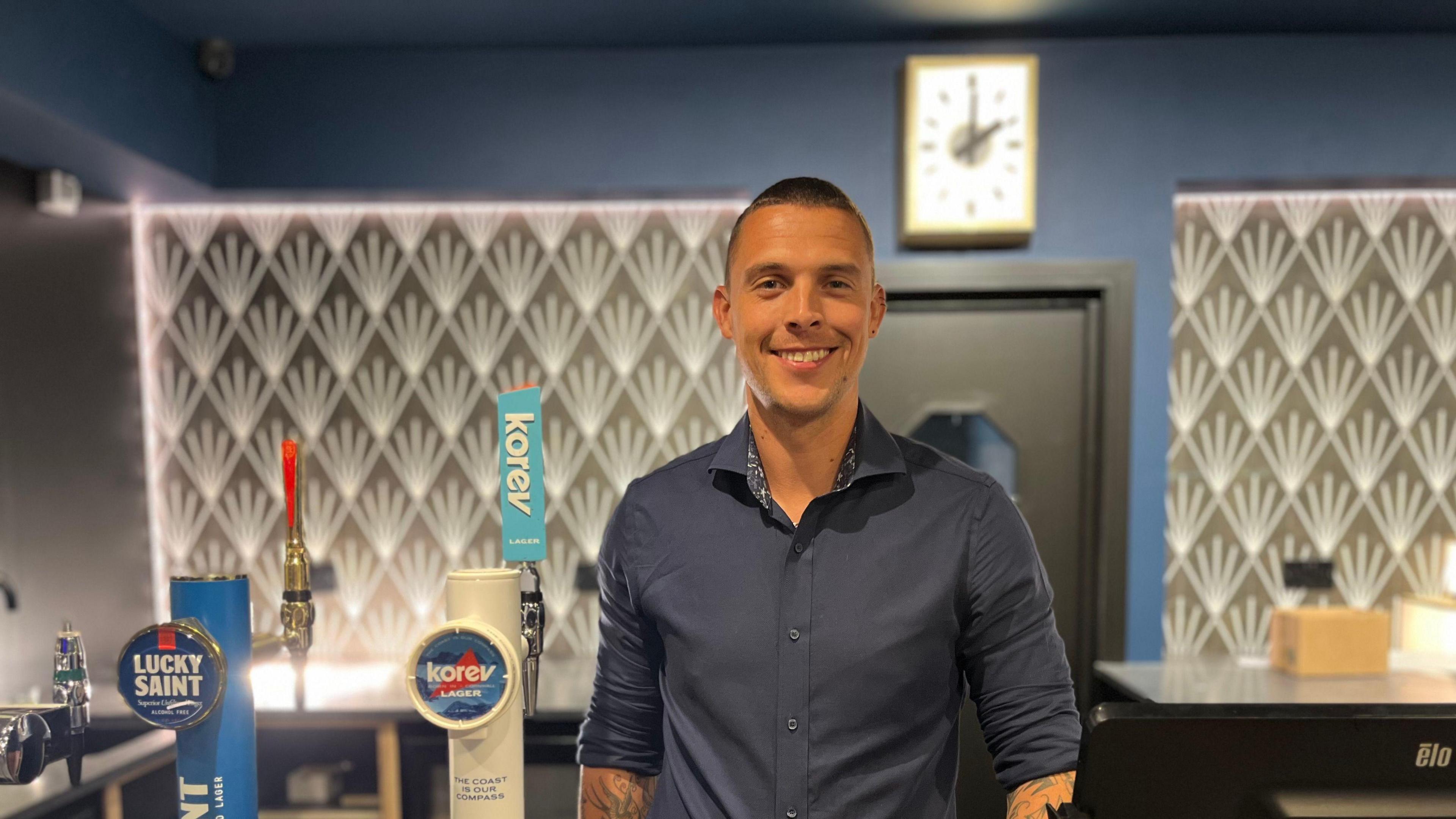 A smiling bar manager who is wearing a dark blue shirt stands behind the bar.
There is a gold patterned 1930s-style wall design on the wall behind him.