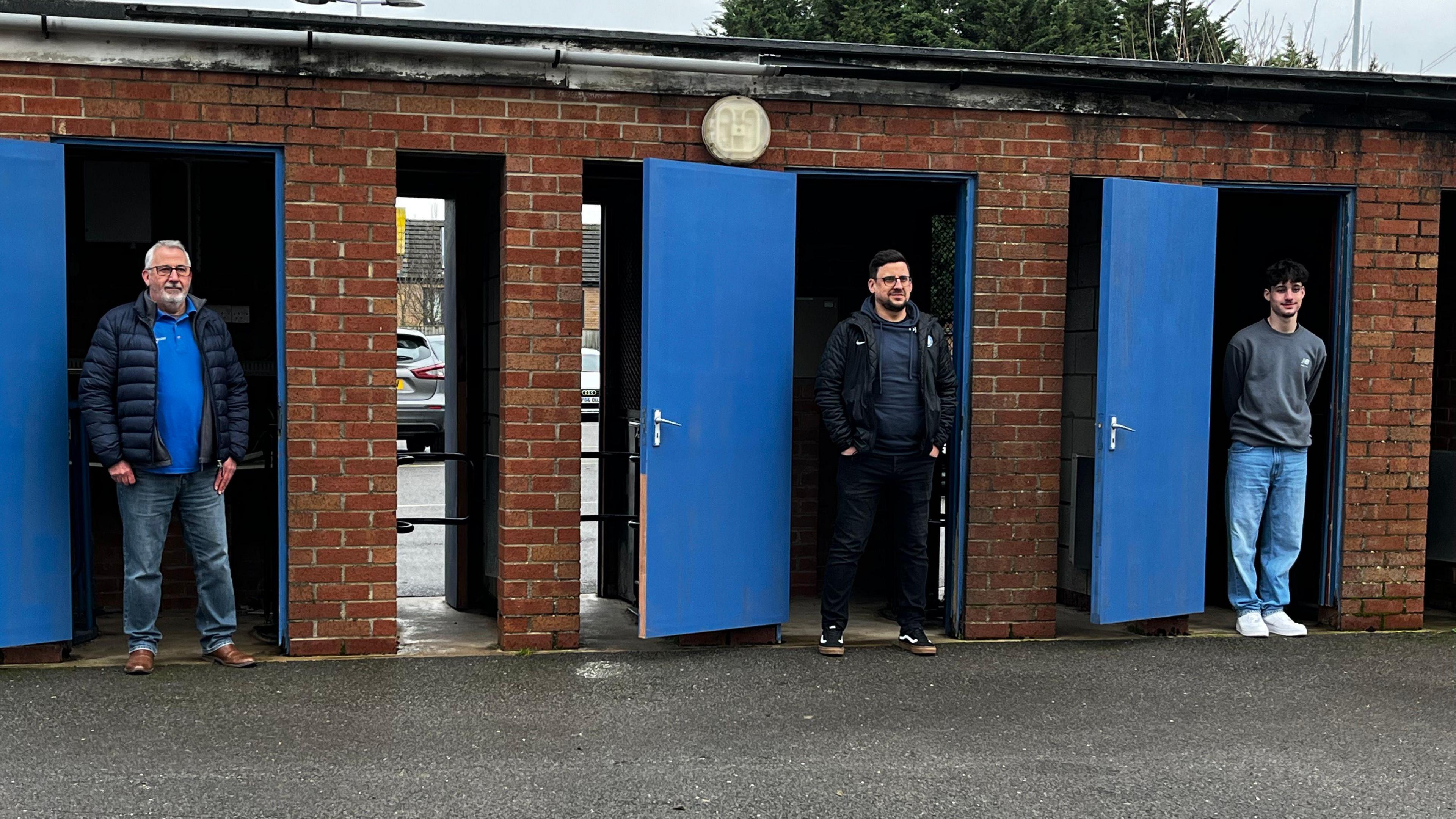 Michael, Ollie and Oscar, are stood in a line. Each one is standing in a different turnstile doorway. The building is outside and red bricked with blue doors, which have been opened. The spaces in which they are stood is where they go to check tickets as fans come into the stadium. 