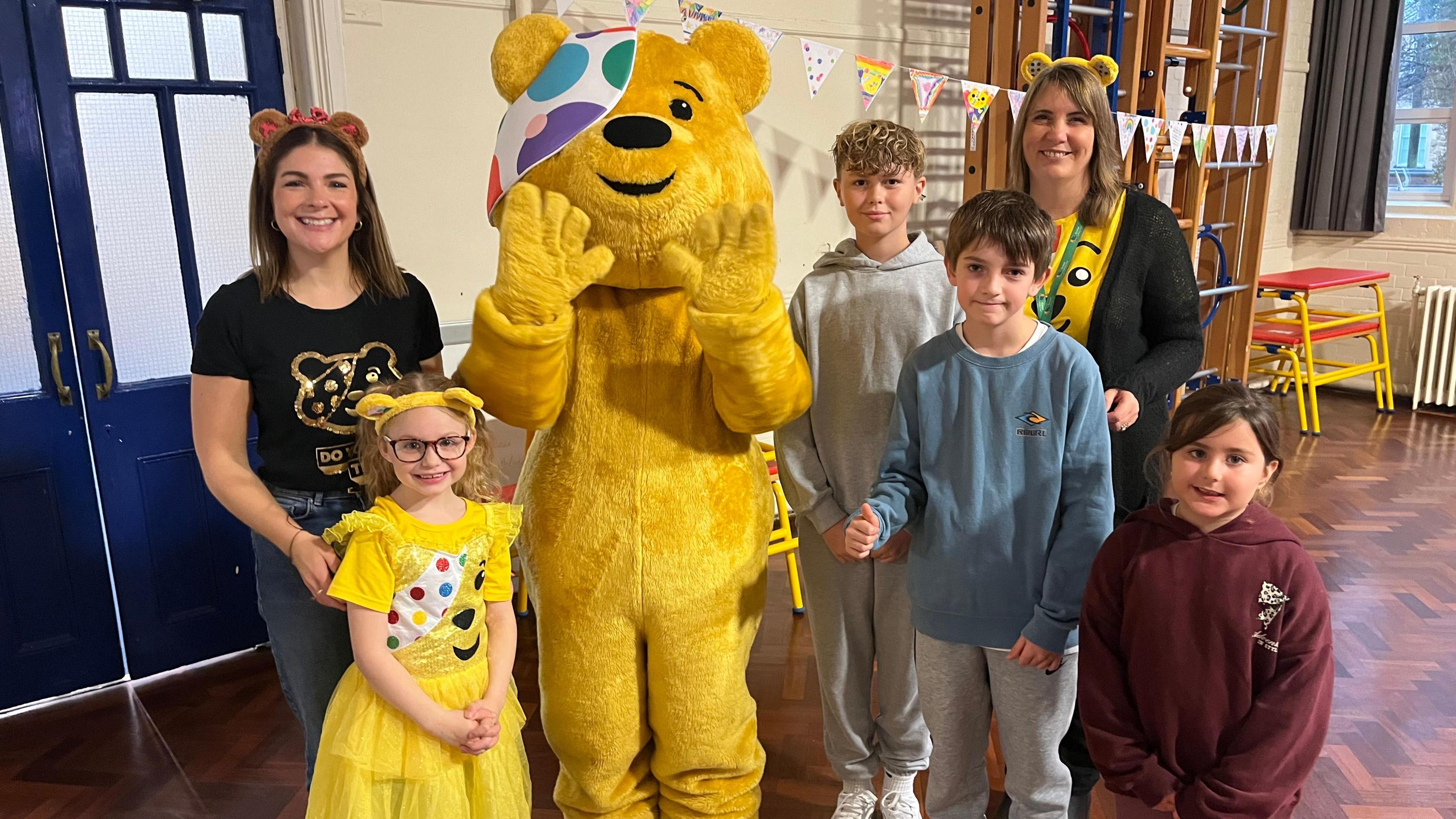 A group of school staff and young students pictured with Pudsey Bear at a primary school. They are all looking at the camera and smiling.