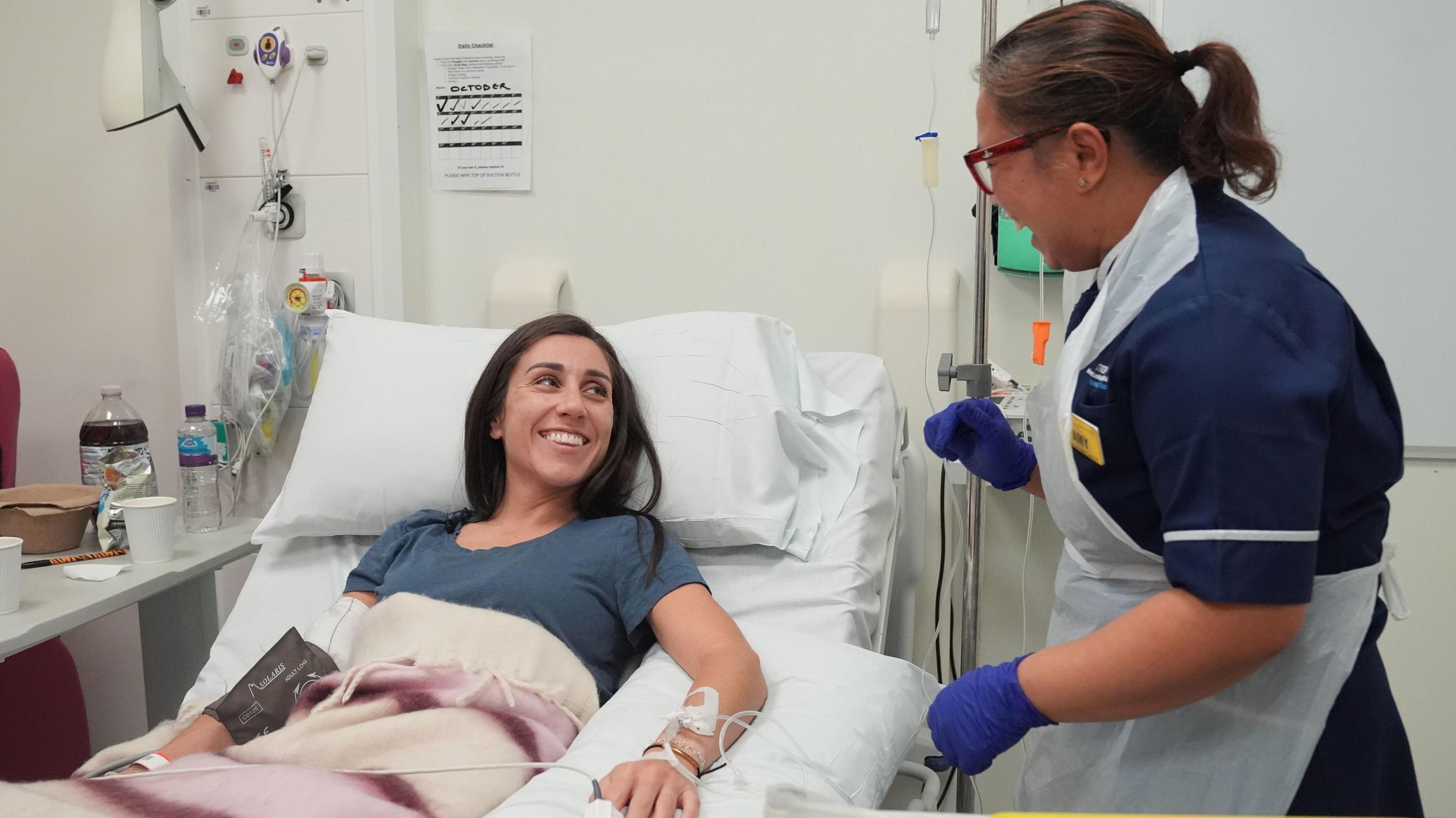 Emily lies in the hospital bed looking up at a female nurse who stands next to her. They are laughing together with wide smiles. The nurse wears blue rubber gloves and a white plastic apron, and has her hair tied back in a ponytail. Lucy has tubes going into her arm and hand, and there is a drip stand next to her bed.