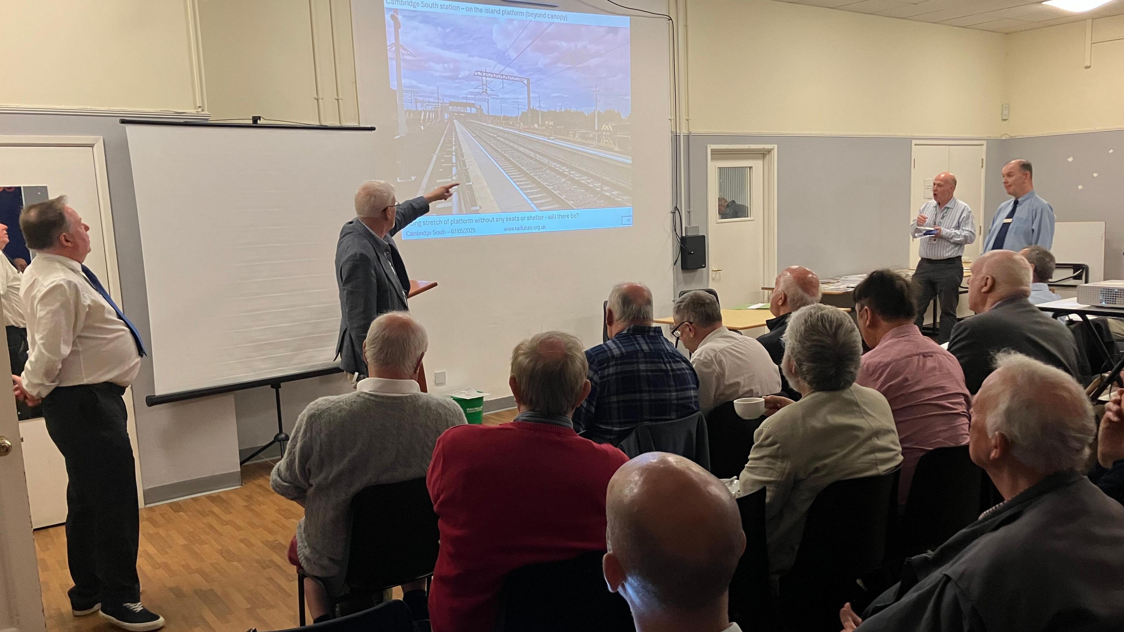 Peter presenting about the station while pointing to the projector- while people facing the slideshow- sitting in rows in the conference room