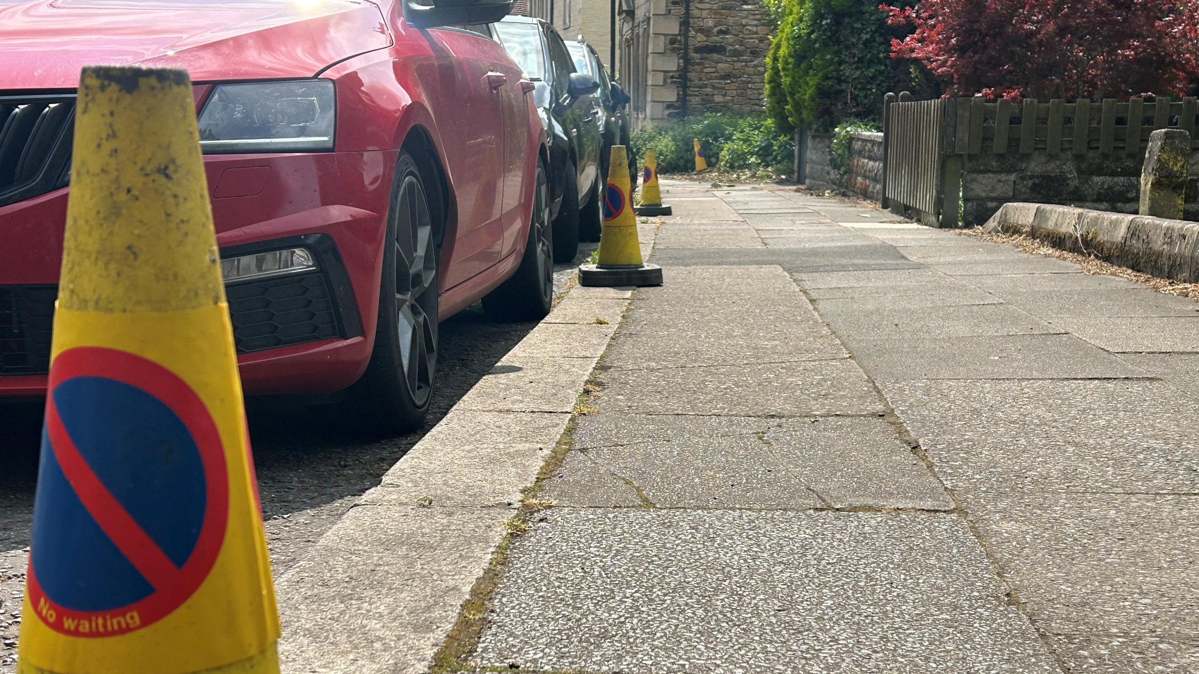 Traffic cones lining a pavement with three cars parked along the kerb.