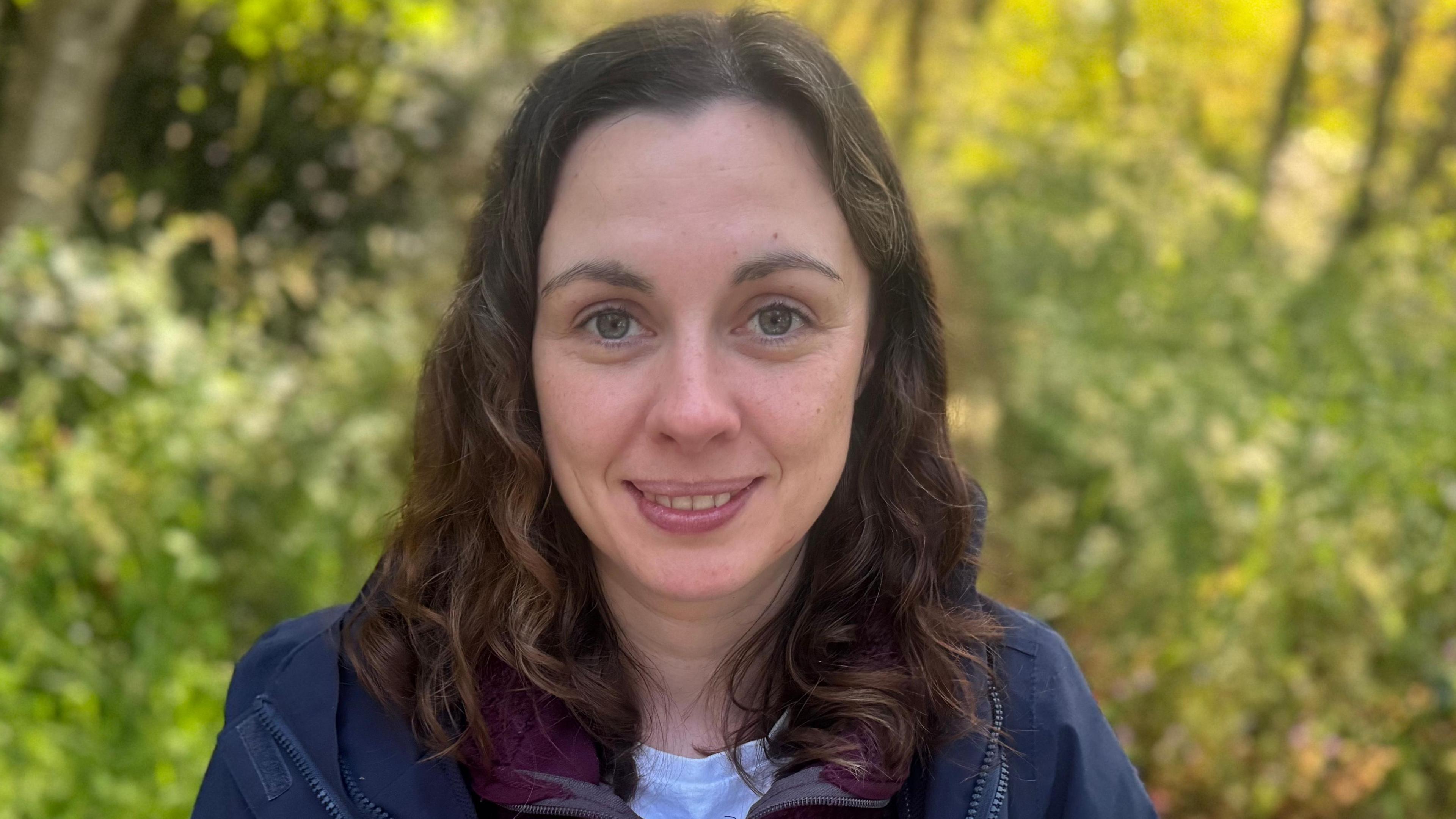 A woman looks directly at the camera. She is smiling and has shoulder-length brown hair. She is wearing a blue jacket and blue t-shirt. She is outside during the daytime, the background is green shrubbery. 