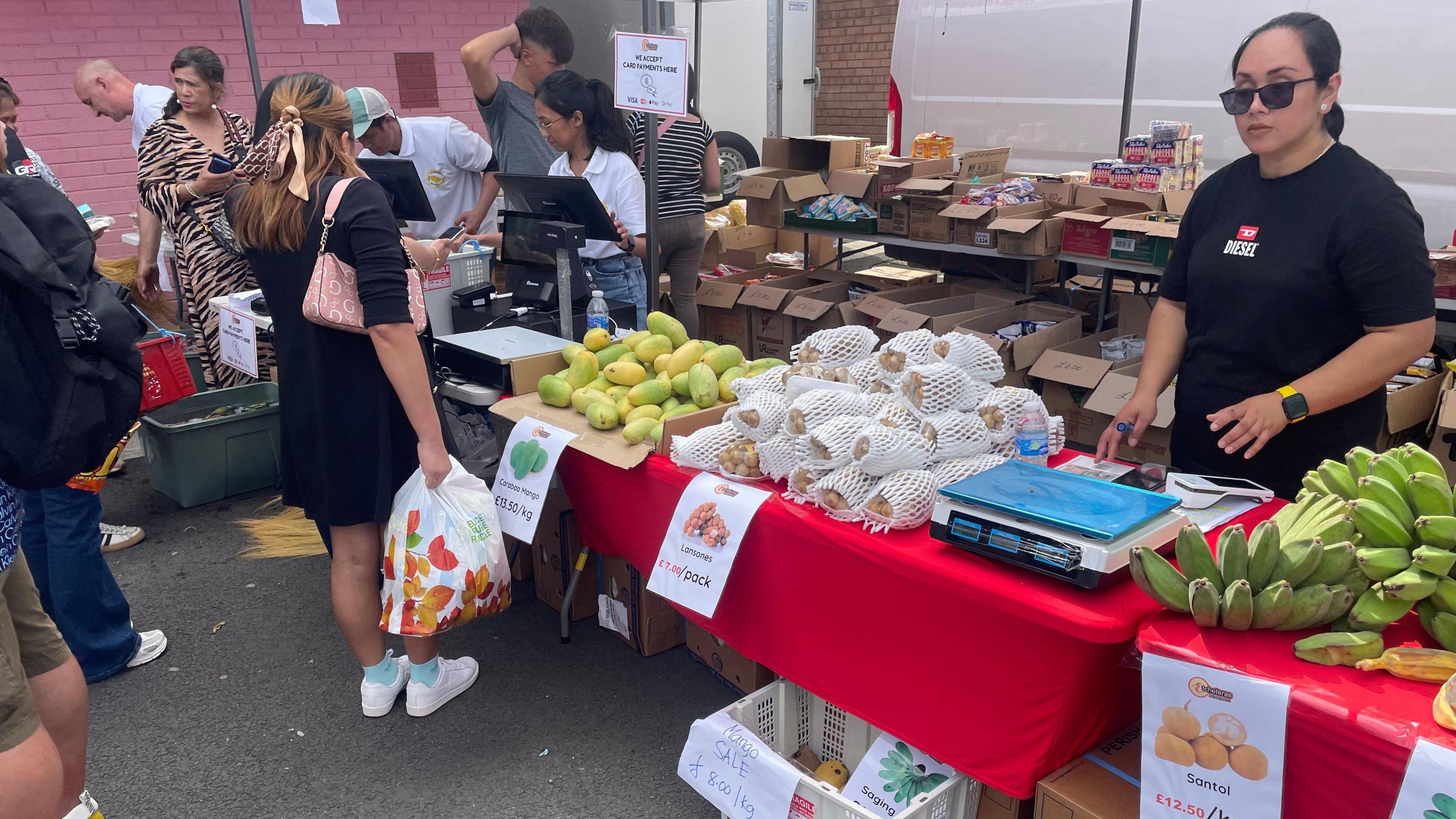 A market stall with fruit on the tables. The tables have a red cloth. Five people are behind the stall whilst a queue of customers wait to be served. Yellow Bananas sit amongst fruit wrapped in a white net, and large round yellow/green fruit. Two scales sit on the table, woman in a black t-shirt and black glasses stands beside one. A woman in a white shirt, brown hair and glasses operates the other one.