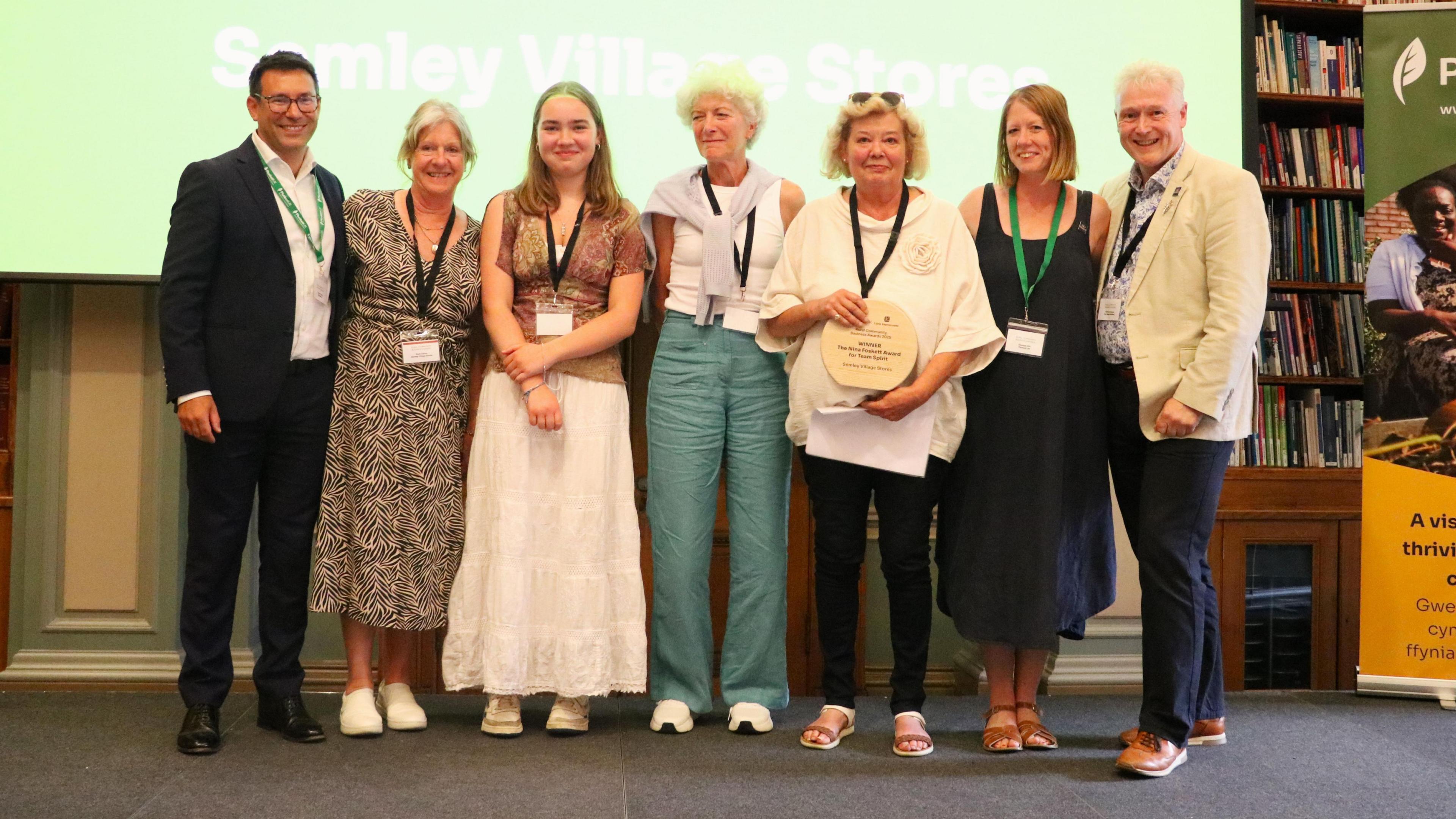 Seven people, two men and five women, stand next to each other on a stage smiling at the camera. The woman third from right holds an award plaque in her hands. They are standing in front of a projection screen, with the word "Semley Village Stores" projected behind them.