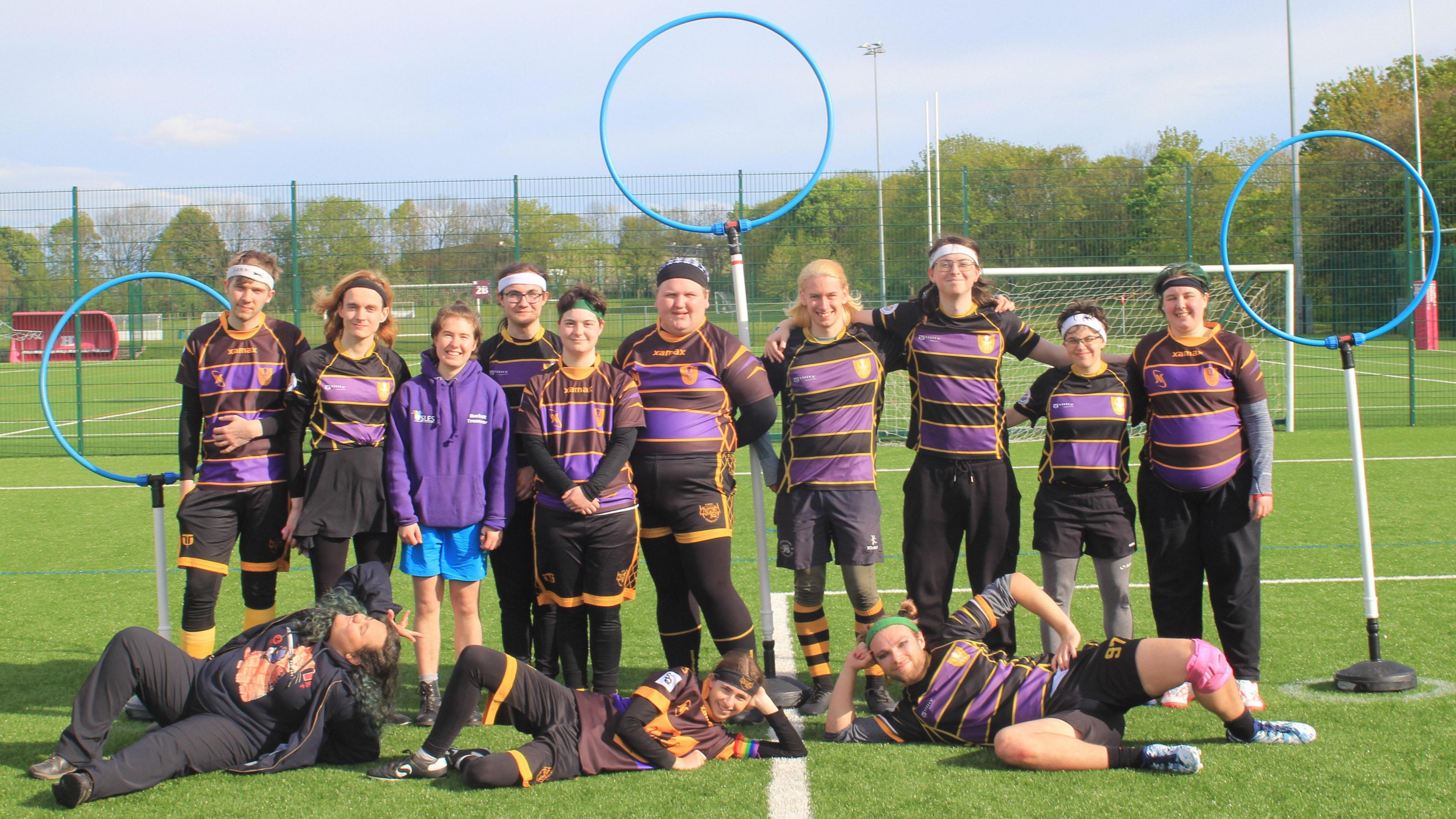 Thirteen people are pictured on a sports pitch. Ten of them are standing arm in arm in front of hoops attached to metal poles. Three players are lying on the floor in front of them.