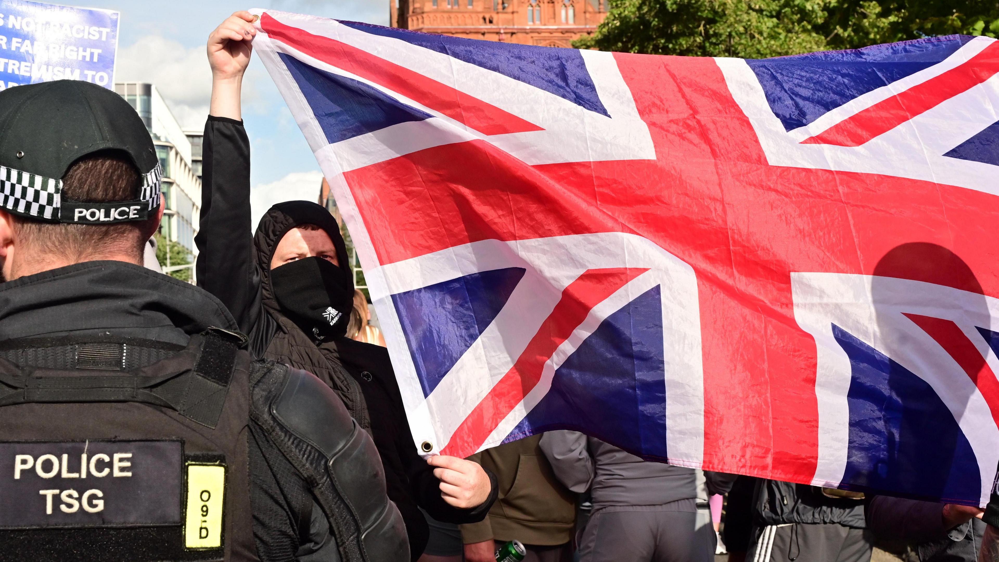 A police officers back and a protester with a mask on holding a union jack