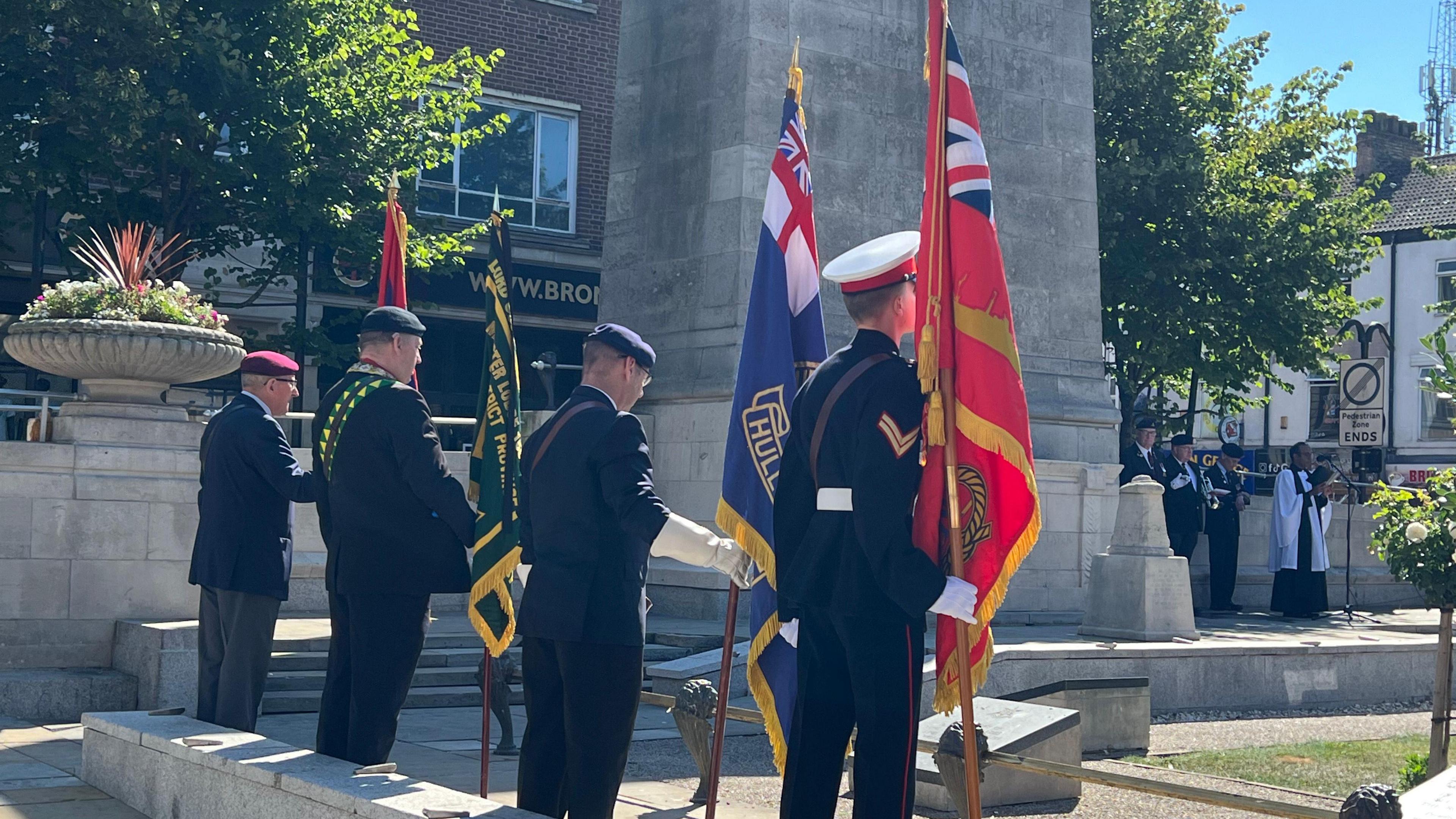 Four military personnel holding flags and standing in a line facing a war memorial. In the background is a band and a person in clerical robes.
