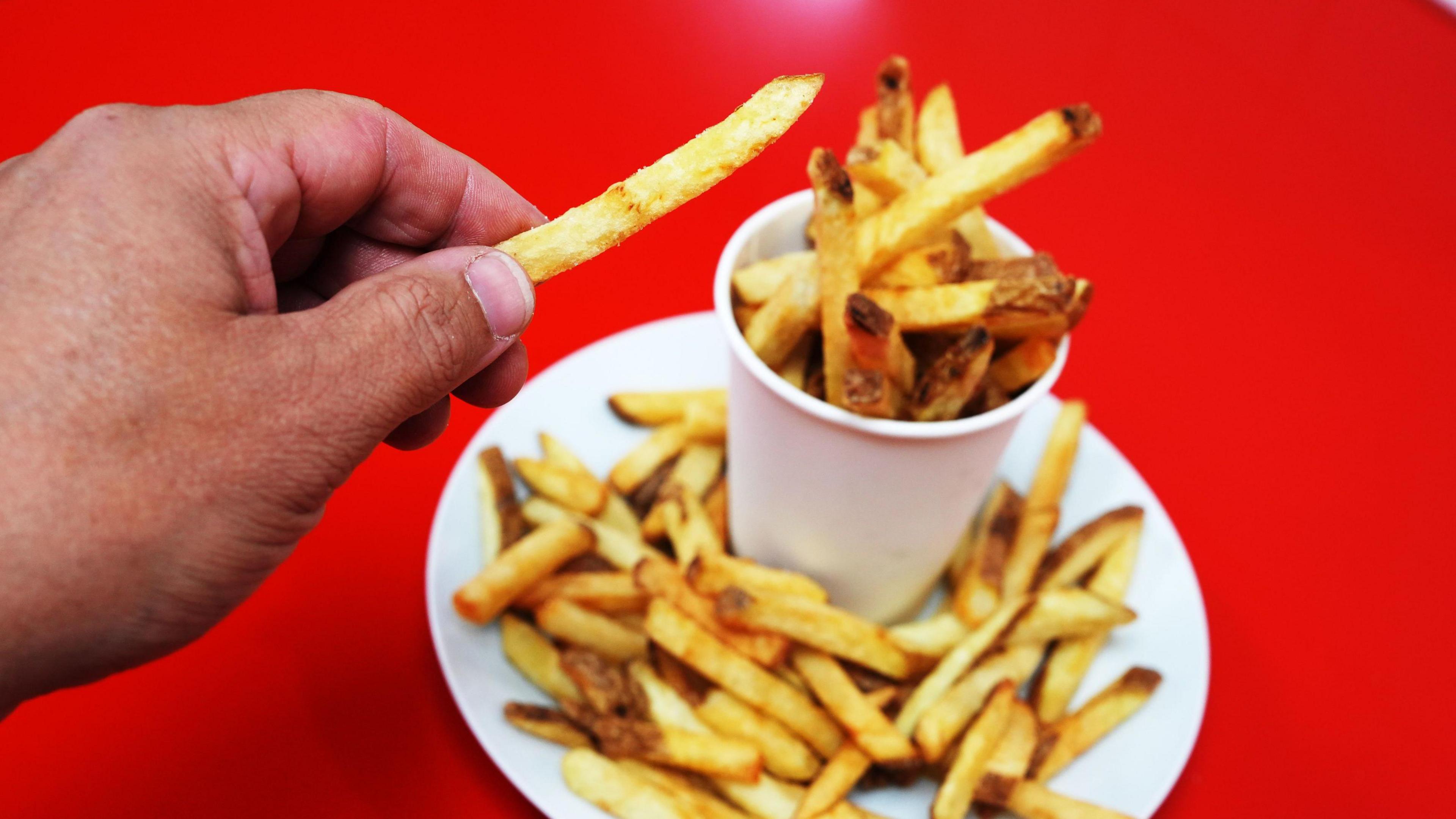 A red background with a white plate and cone of French fries on top. A persons hand is in view. They are taking a french fry out of the pot and holding it towards the camera.