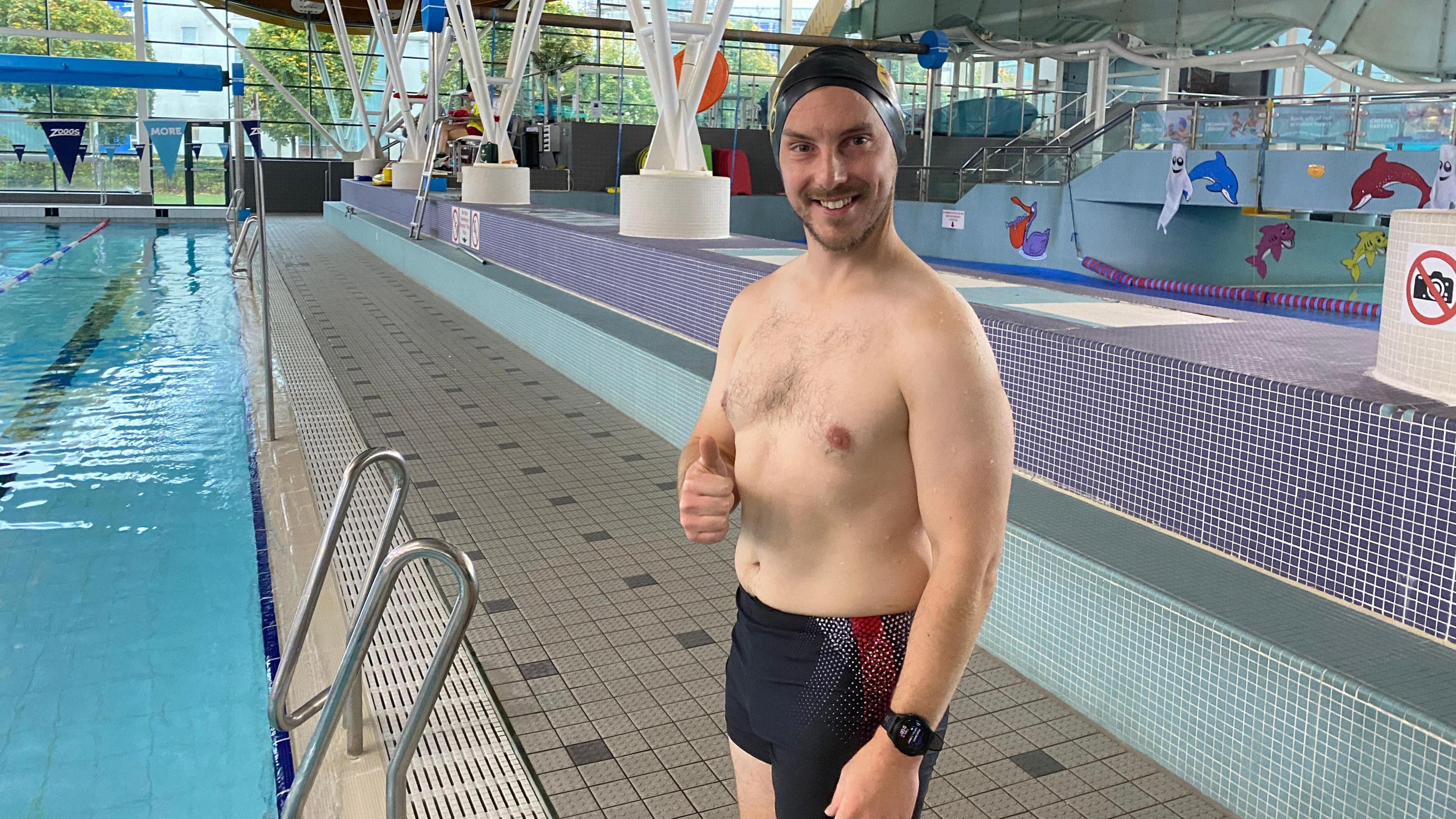 Chris stands by the pool with his swim cap on which has the Thousand Mile Challenge logo on it. He has dark blue swimming trunks and he's smiling at the camera and giving it the thumbs up. There is a pool slide behind him.