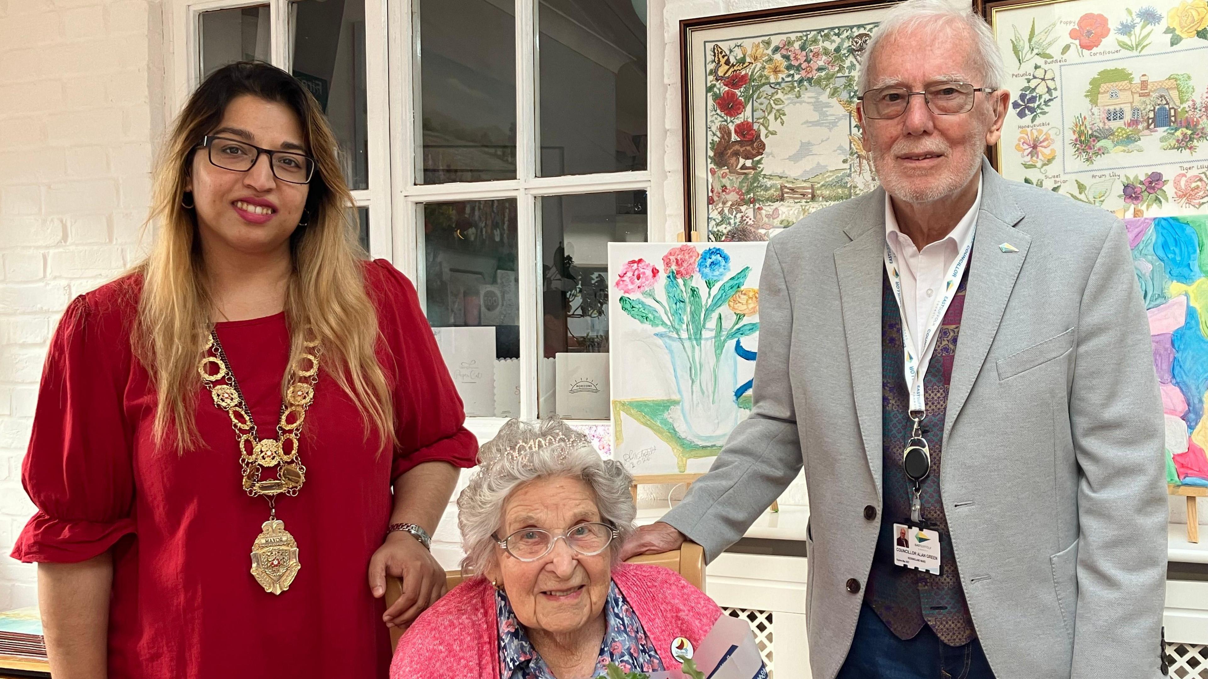 The Mayor of Lowestoft Nasima Begum, wearing her ceremonial chain of office stands beside a councillor Alan Green representing East Suffolk Council wearing a grey jacket. Seated in the middle is Olive wearing a tiara in her hair and a pink cardigan.  