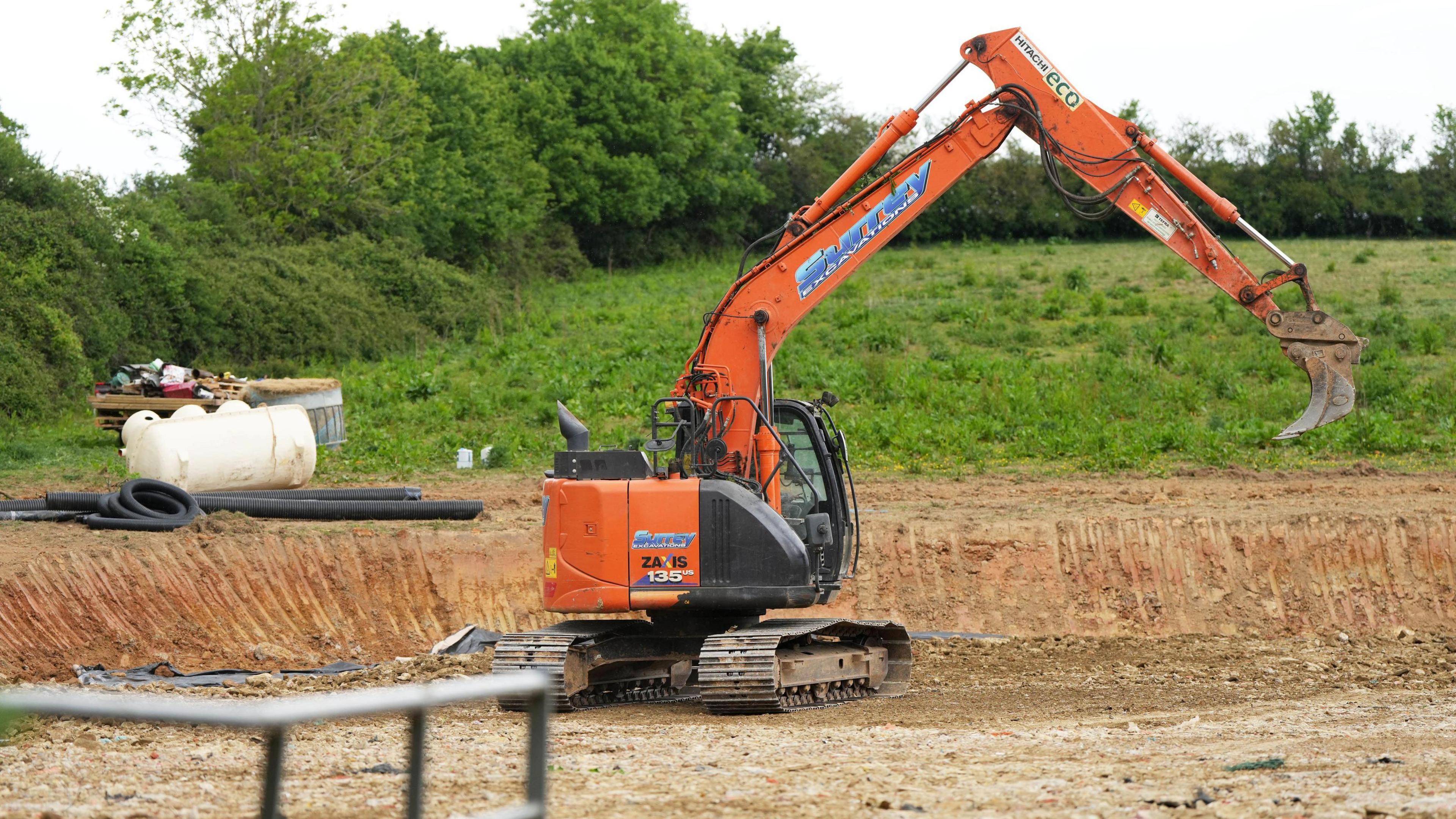 An orange digger in a field with grass and dirt in. A white septic tank also sits in the field.