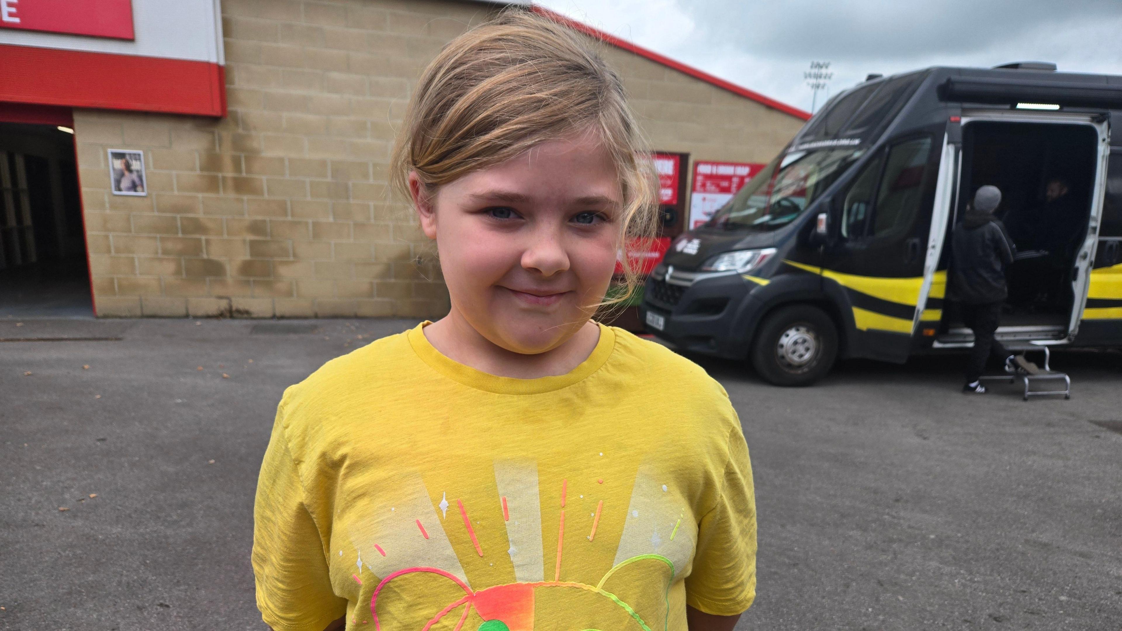Mia wearing a yellow t-shirt with an image of Pudsey on it. She has her hair tied up and her hands behind her back smiling at the camera. She is standing outside and there is a black van behind her with people in the doorway of it.