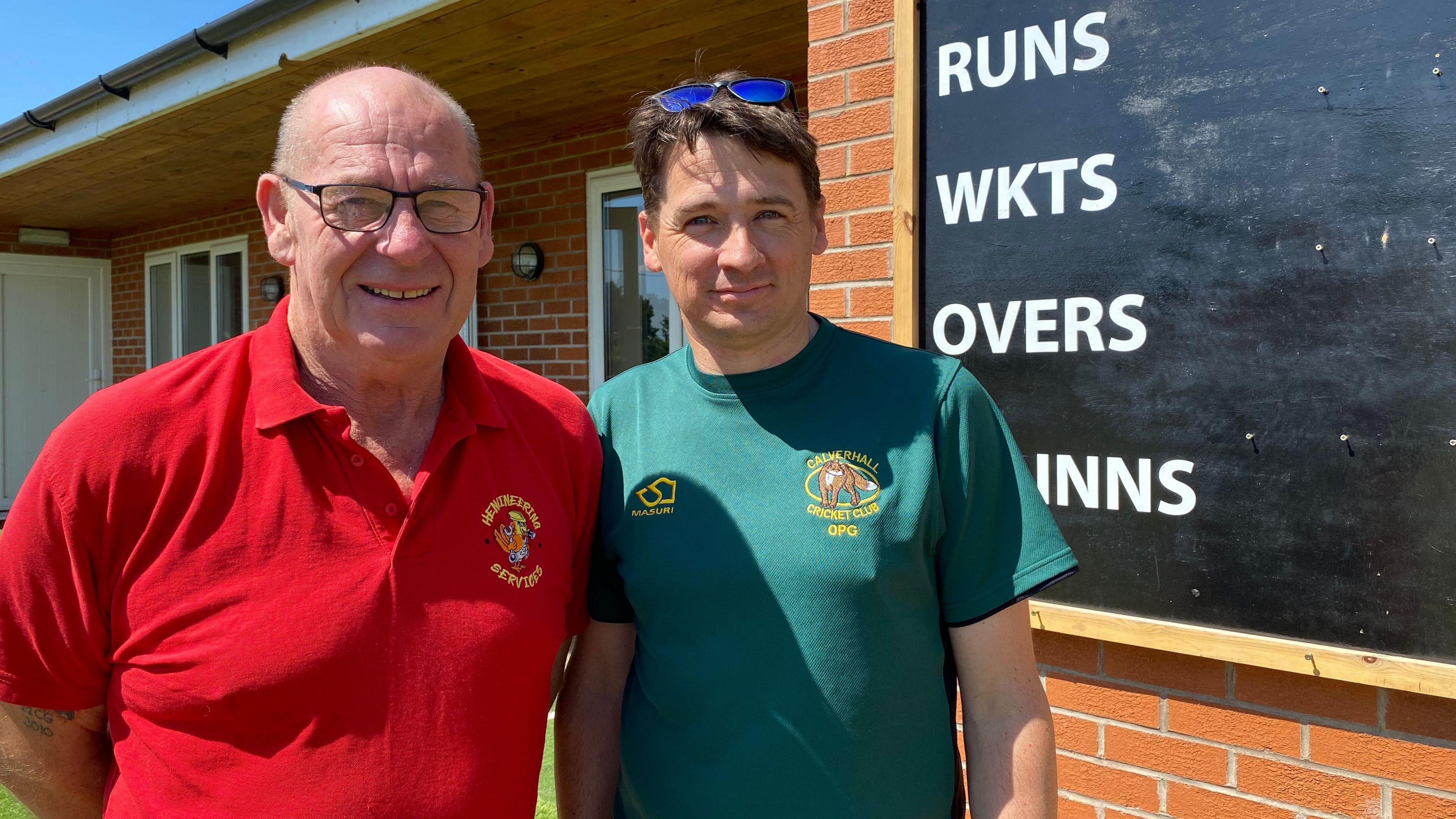 A man with very short grey hair and wearing black glasses and a red T-shirt, is standing next to a man with sunglasses perched on top of his head. He has short brown hair and and is wearing a dark green-coloured T-shirt. They are both standing outside a cricket club and behind them is a blackboard with white writing that reads "RUNS, WICKETS, OVERS and 1st innings".