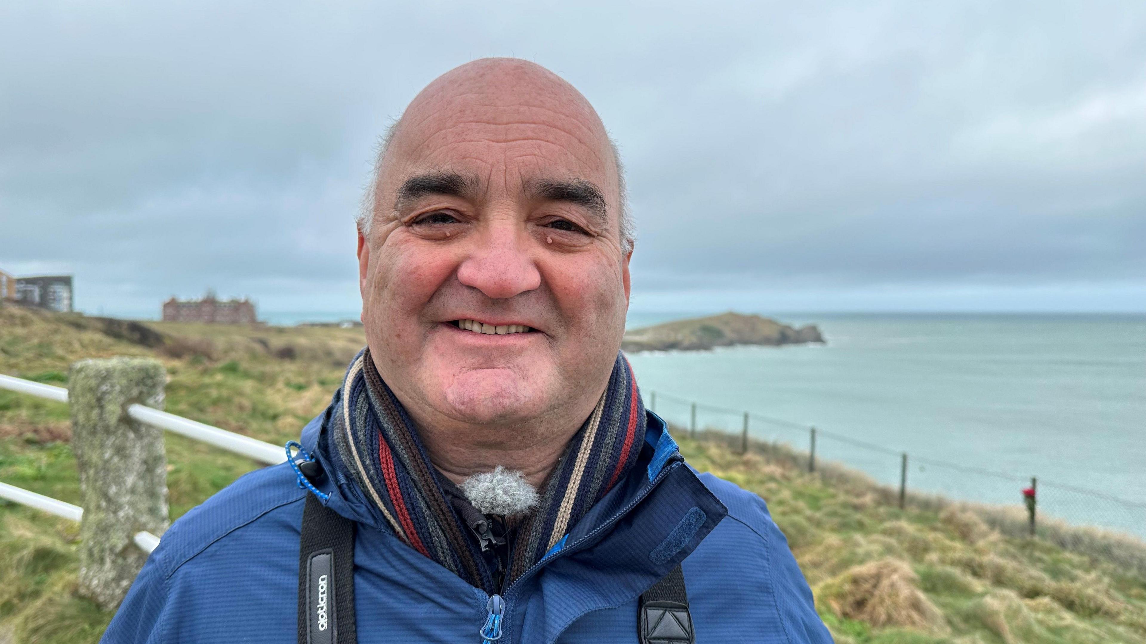 A man wearing a blue raincoat smiling. He is stood on the coast in front of the sea with grass and a fence just behind him