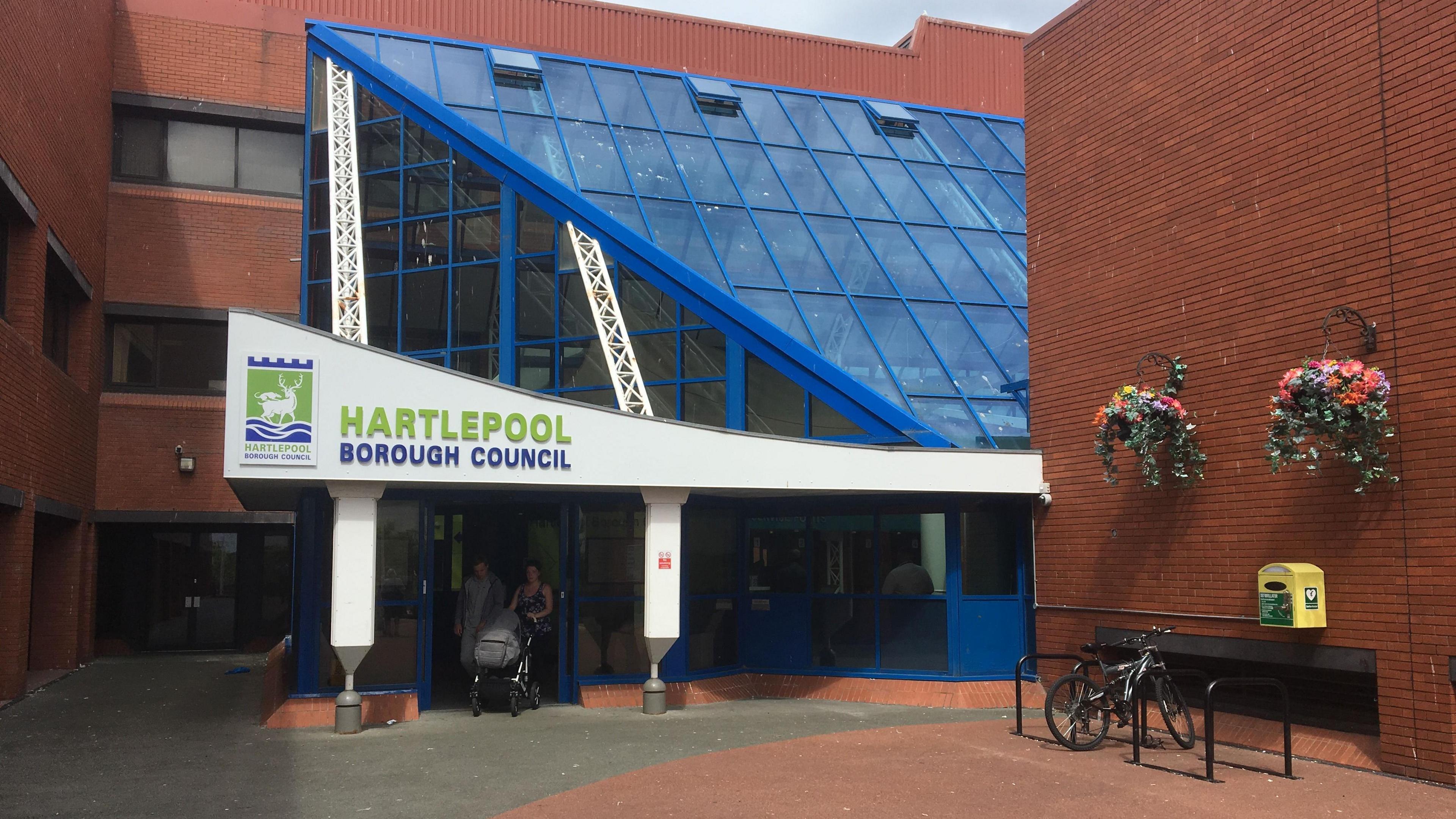 Picture of the entrance to Hartlepool Borough Council. A woman pushing a baby pram is exiting the blue, glass-fronted building with a man. There is a bike rank on the right with two hanging baskets of flowers above.