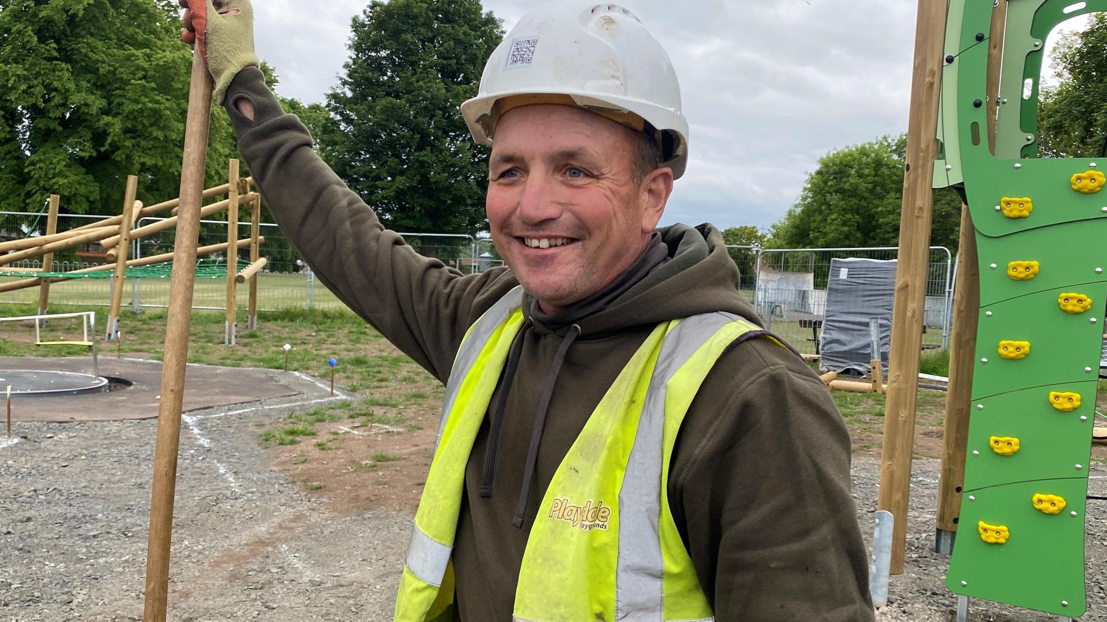 Steven Parkinson is standing in the middle of the playground in Scotts Park, holding a long rake in one hand and smiling He is about 40 years old and wearing a white safety helmet and a yellow hi-vis jacket. Behind him there are items of play equipment, closed off by temporary metal fences. 