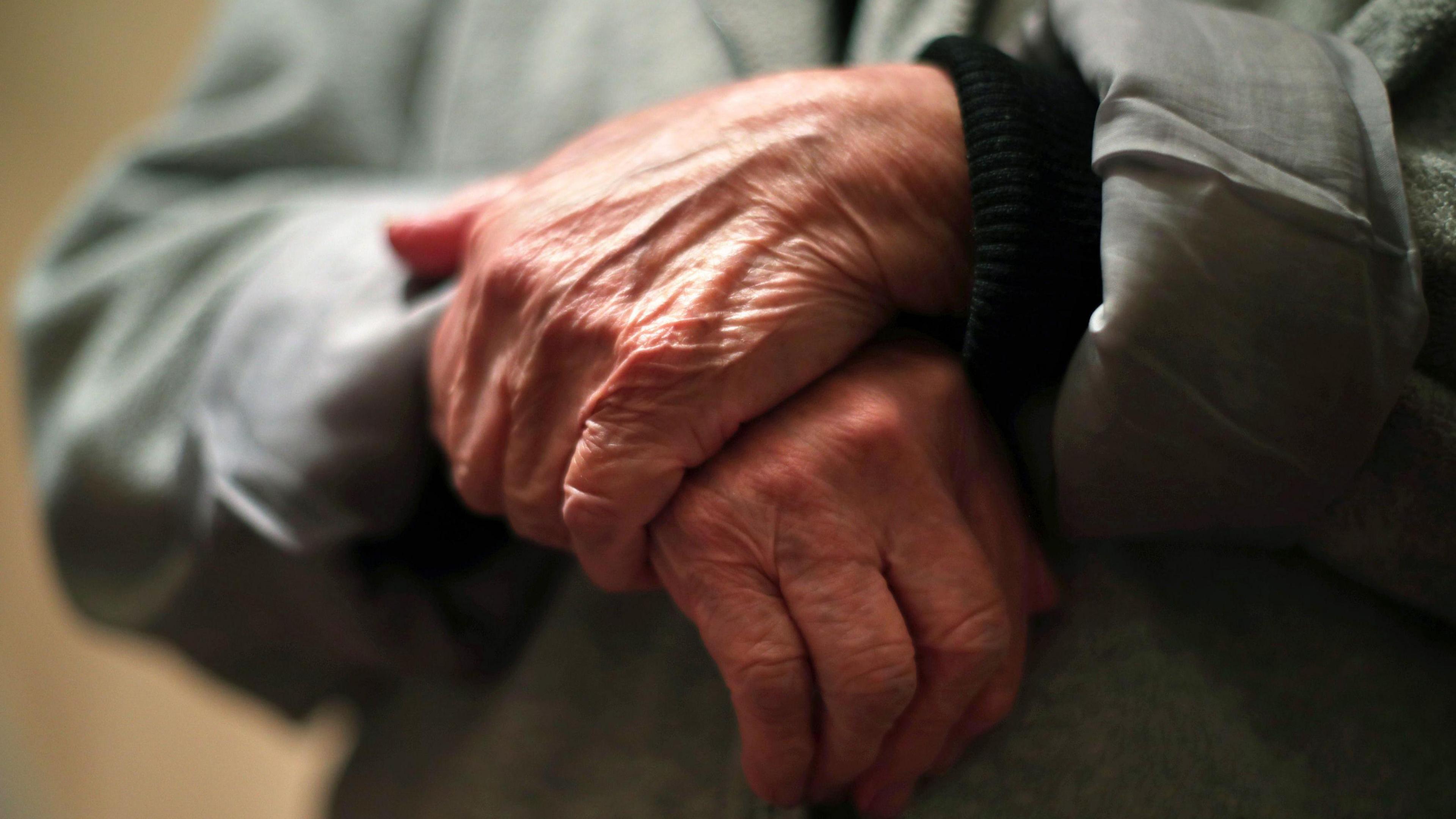 A close-up image of the hands of an elderly person, they are crossed over in front of their body. Their arms are covered with the sleeves of a grey jacket, which is slightly out of focus. A beige-coloured wall is in the background.