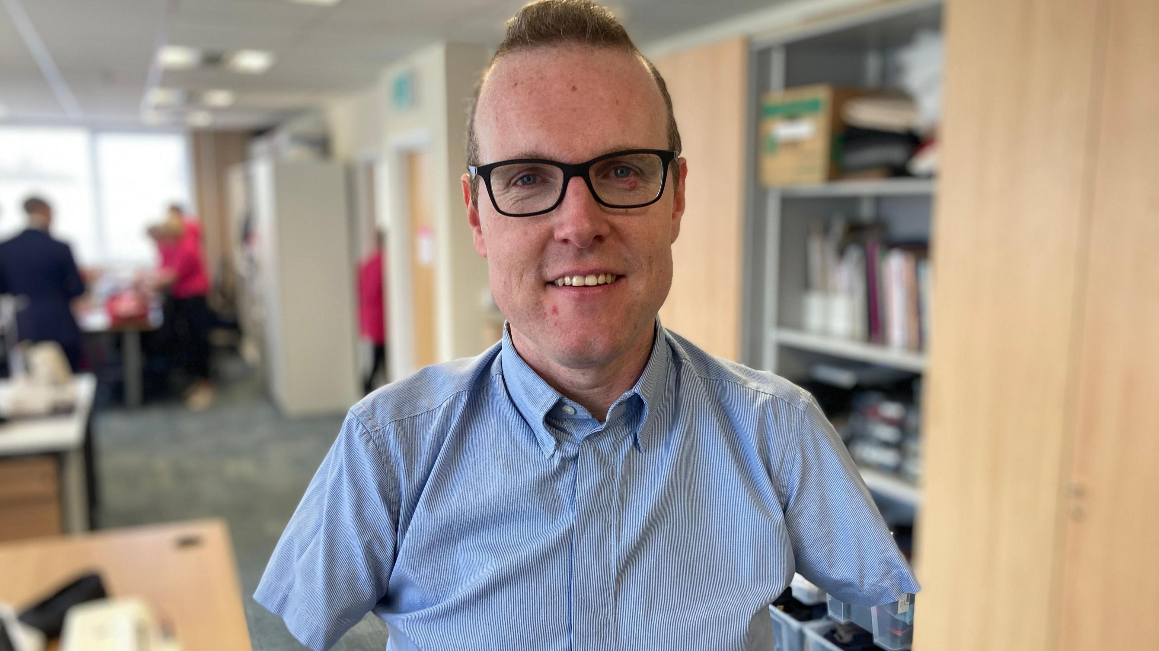 Andy in an office, wearing a blue short-sleeved shirt and black-rimmed glasses. He has closely cropped fair hair. Andy has very short arms and no hands.