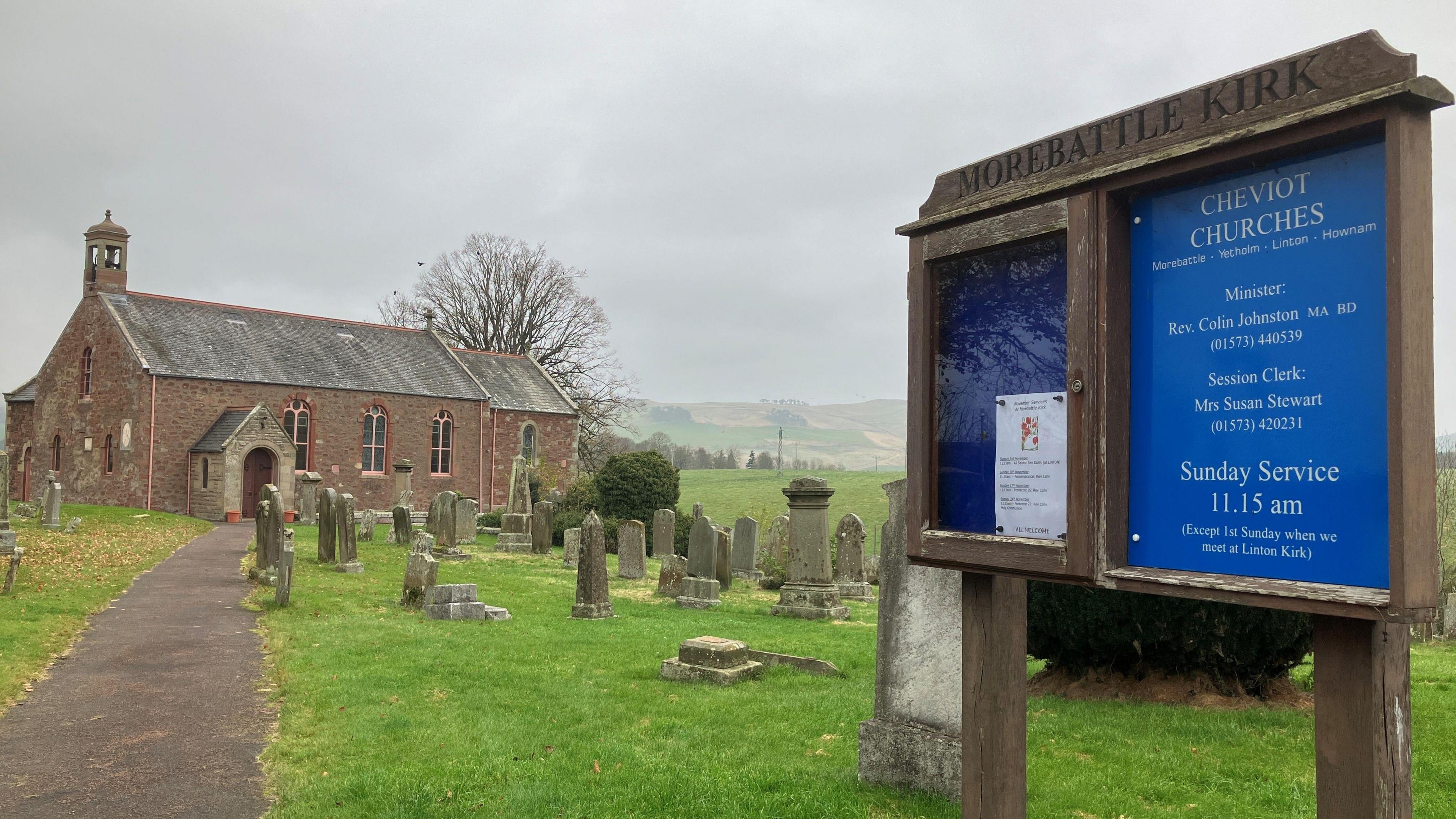 A notice board and sign for Morebattle Kirk stands in front of a graveyard and old church building in the Borders countryside