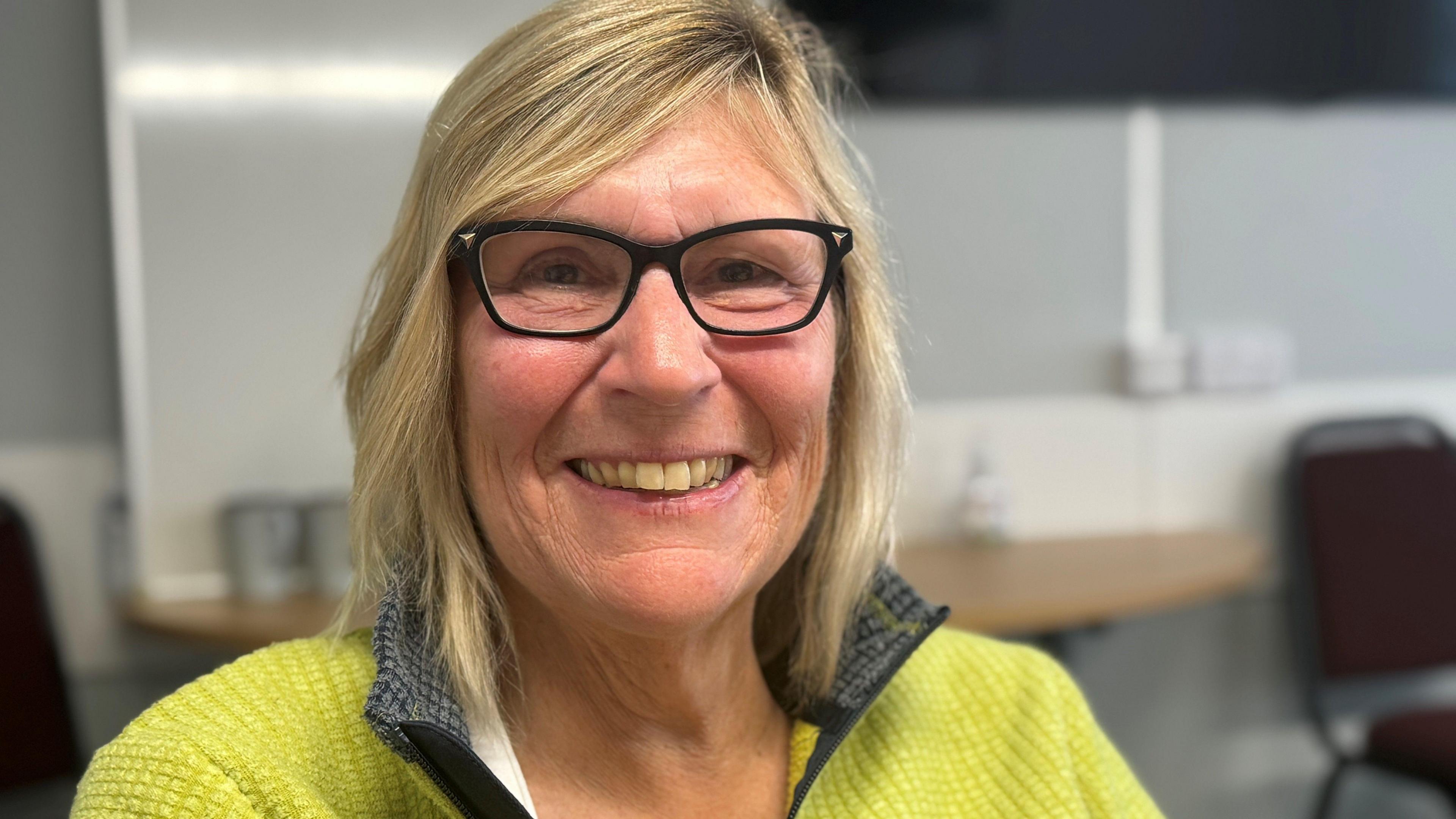 A woman with shoulder-length blonde hair and black glasses is smiling at the camera. She is wearing a green jumper. There is a grey wall and dark red chair behind her.