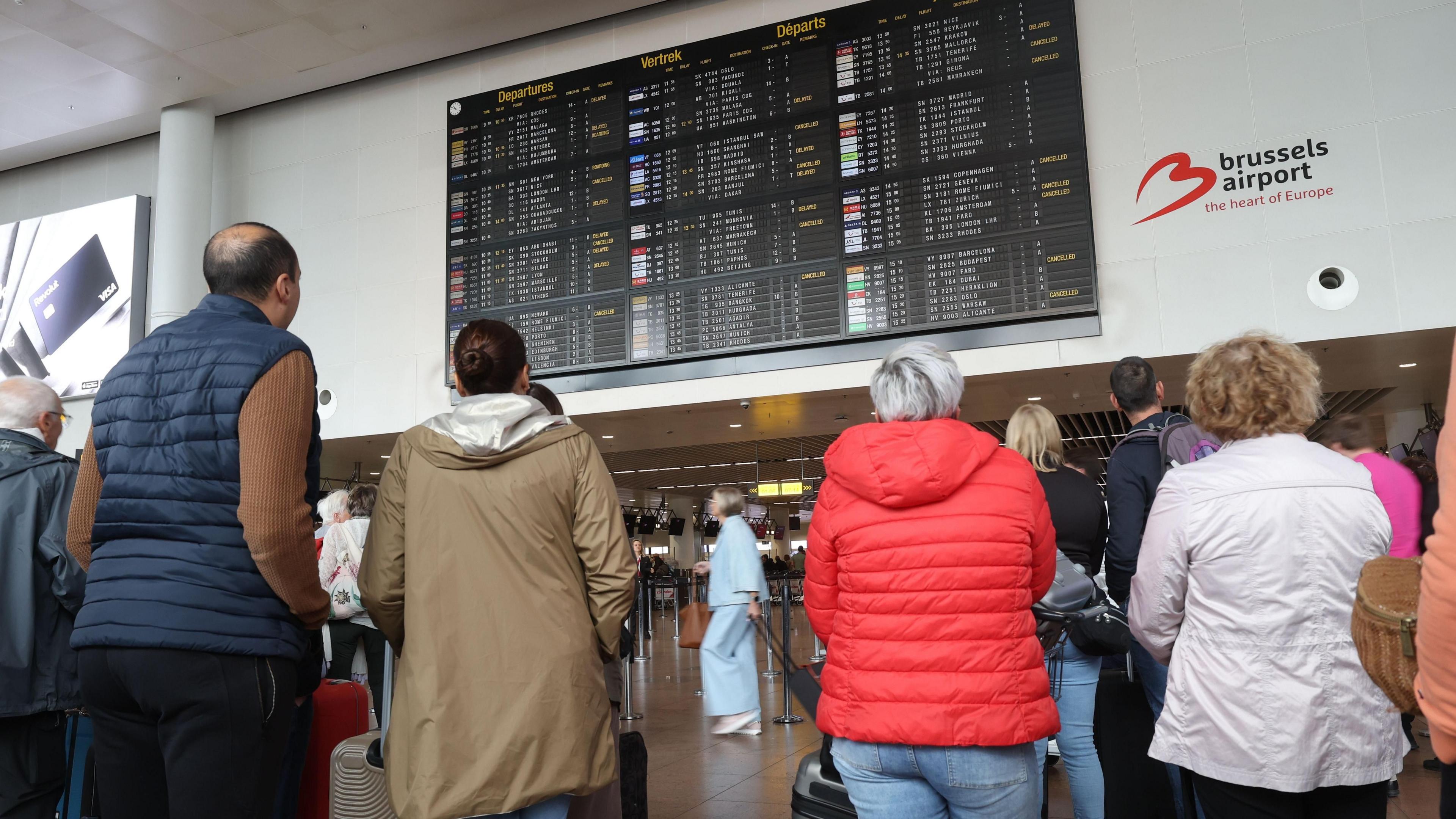 A group of people leaning on wheeled suitcases look up at an airport departures board. In front of them are a series of airport check-in desks.