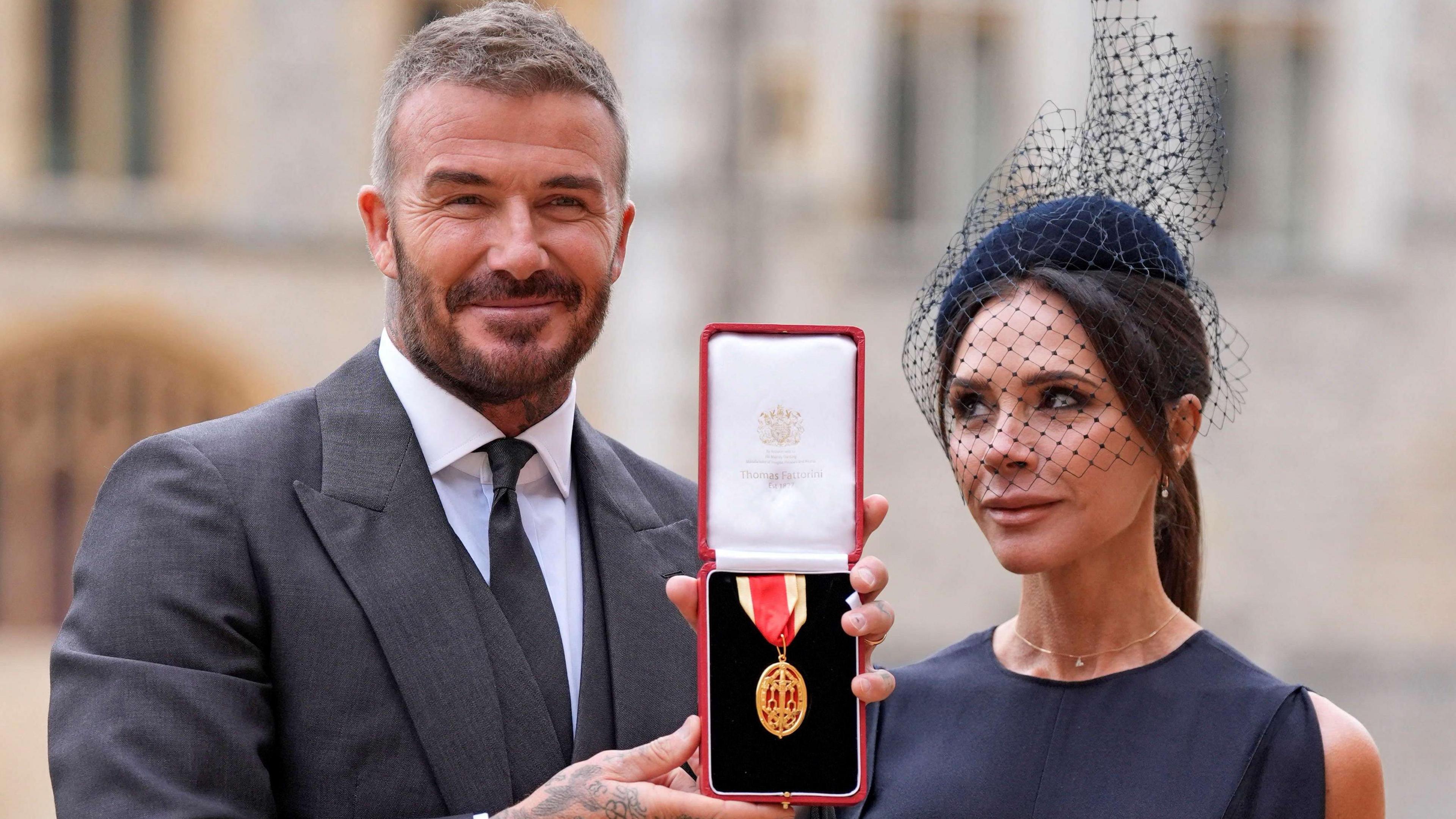 Sir David and Lady Beckham pose for the cameras outside Windsor Castle with Sir David's honour. He is wearing a grey suit, white shirt and grey tie and she is in a navy sleeveless dress and pillarbox hat with netting.