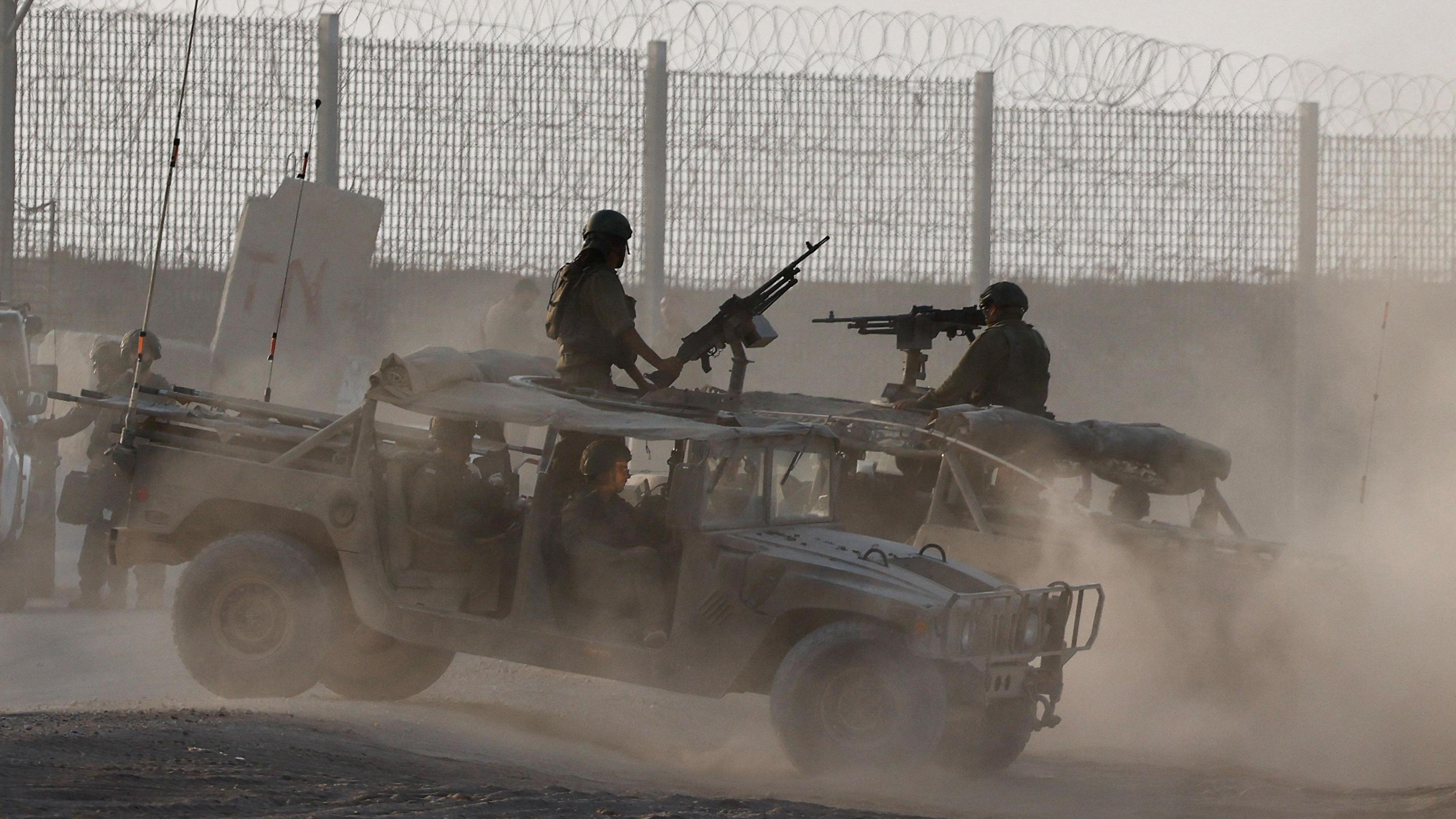 Members of the Israel Defence Force stand in two open-topped military vehicles near a fenced border, which is topped with barbed wire. Each vehicle has a large gun extending from its roof, manned by a person in a helmet and camoflague uniform.