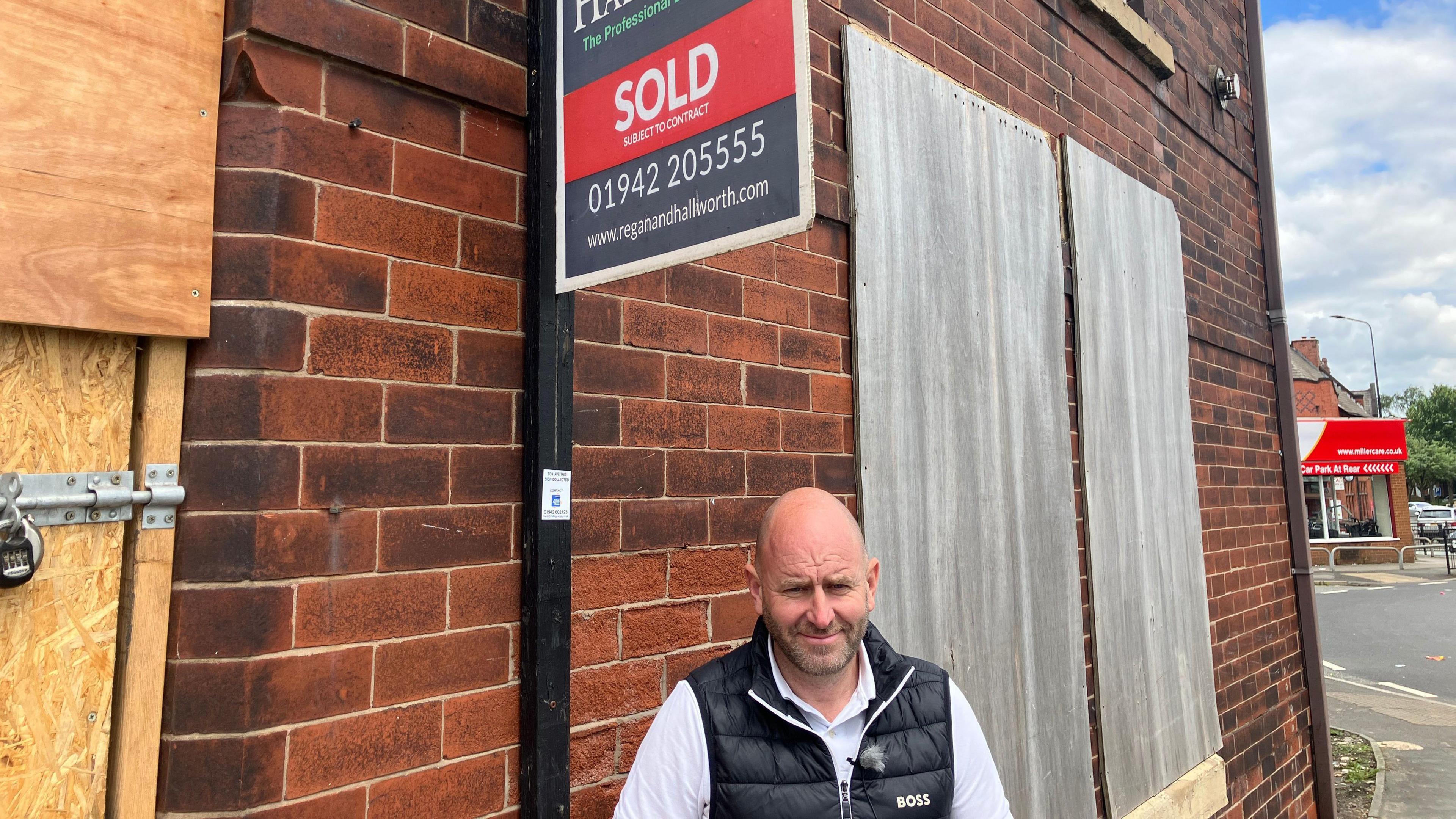 Andy Lyon standing in front of a boarded up property below a for sale sign he has bought