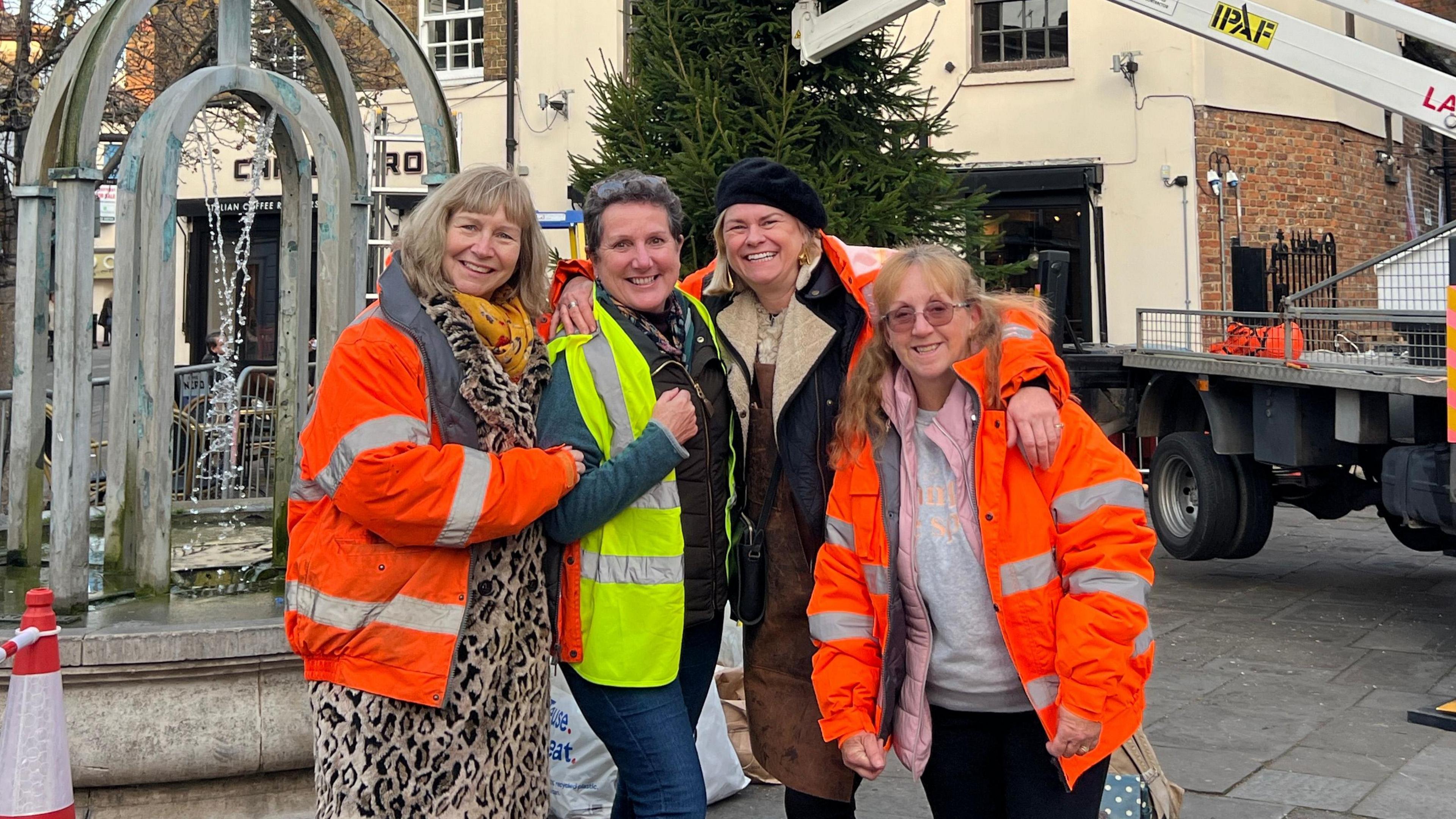 Four women, smiling and looking straight at the camera, in the middle of Hertford. Three are wearing high-vis jackets, and one woman in the middle has a black beret on. A Christmas tree is behind them and a lorry is putting it in place. You can see shops behind and a monument. 