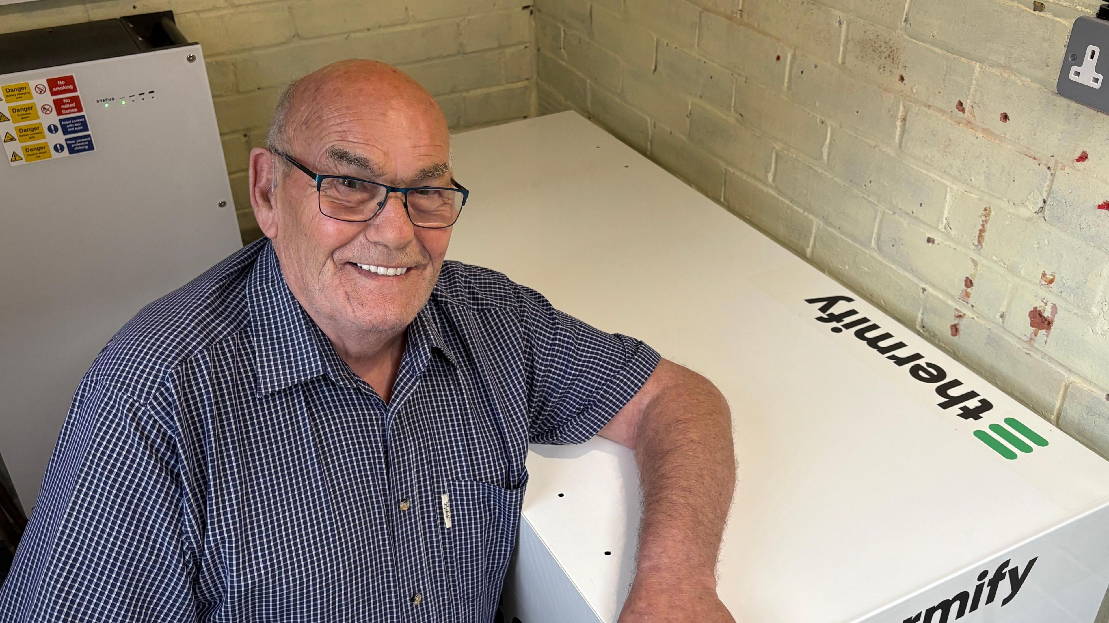 Terrence Bridges kneeling down and smiling on the left of the frame, while leaning an arm over a large white object with "Thermify" written on it, which is on the right of the image. Terry is wearing a short-sleeved blue-checked shirt, which is open at the neck, and black-rimmed rectangular glasses. He is inside a small room, with brick walls. The bricks have been painted a cream colour. An unused plug socket is just visible in the top right of frame. Behind Terrence is another piece of electrical equipment with a white front, several warning stickers and two green lights.