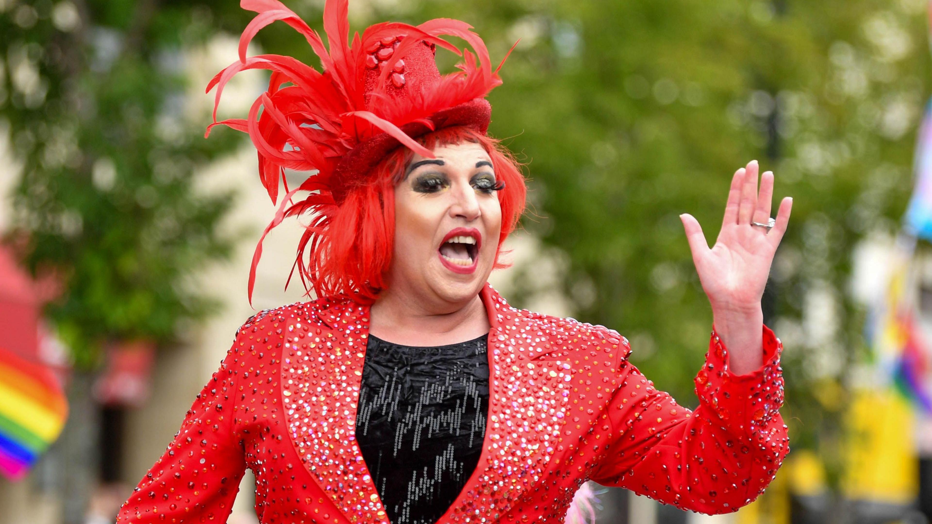 A person on top a float at a parade. They're wearing a sparkly red jacket and a black top and a red top hat with feathers.