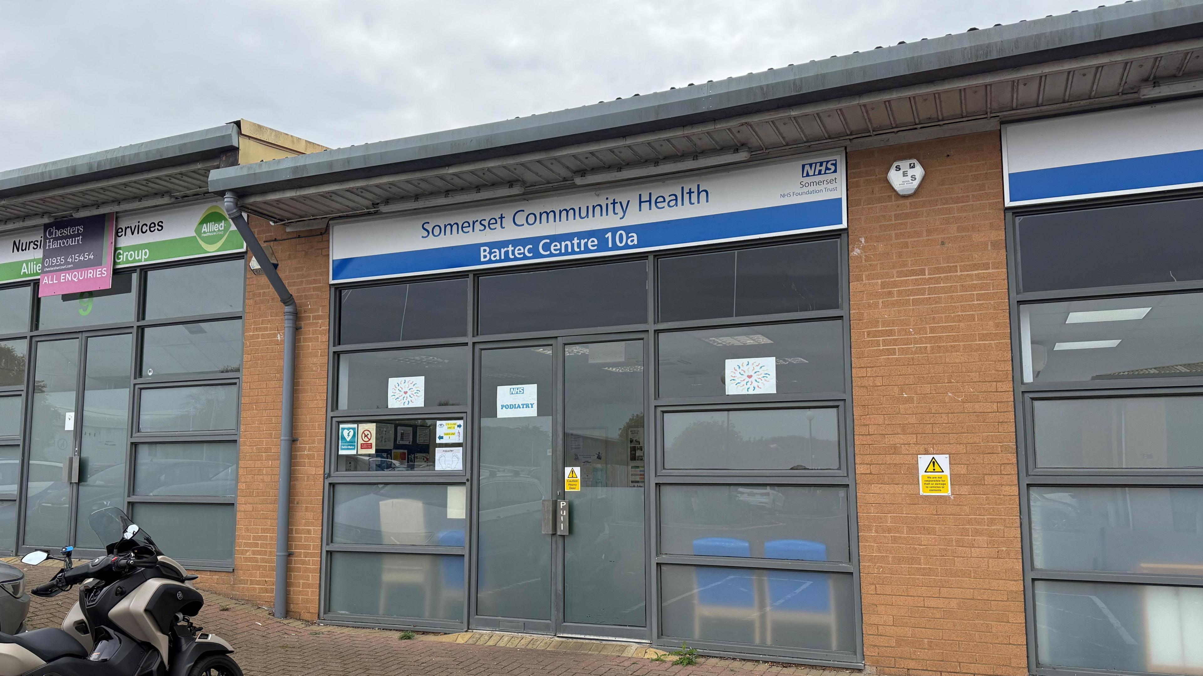 Building in an industrial estate with a "Somerset Community Health" sign with an NHS Somerset logo. There's big windows and a large door. It has a sign that says "Podiatry".